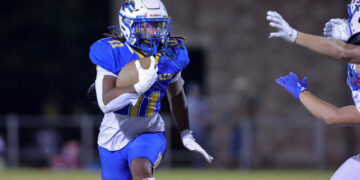 American football player in blue and yellow jersey runs with the ball, evading a defender reaching in from the right.