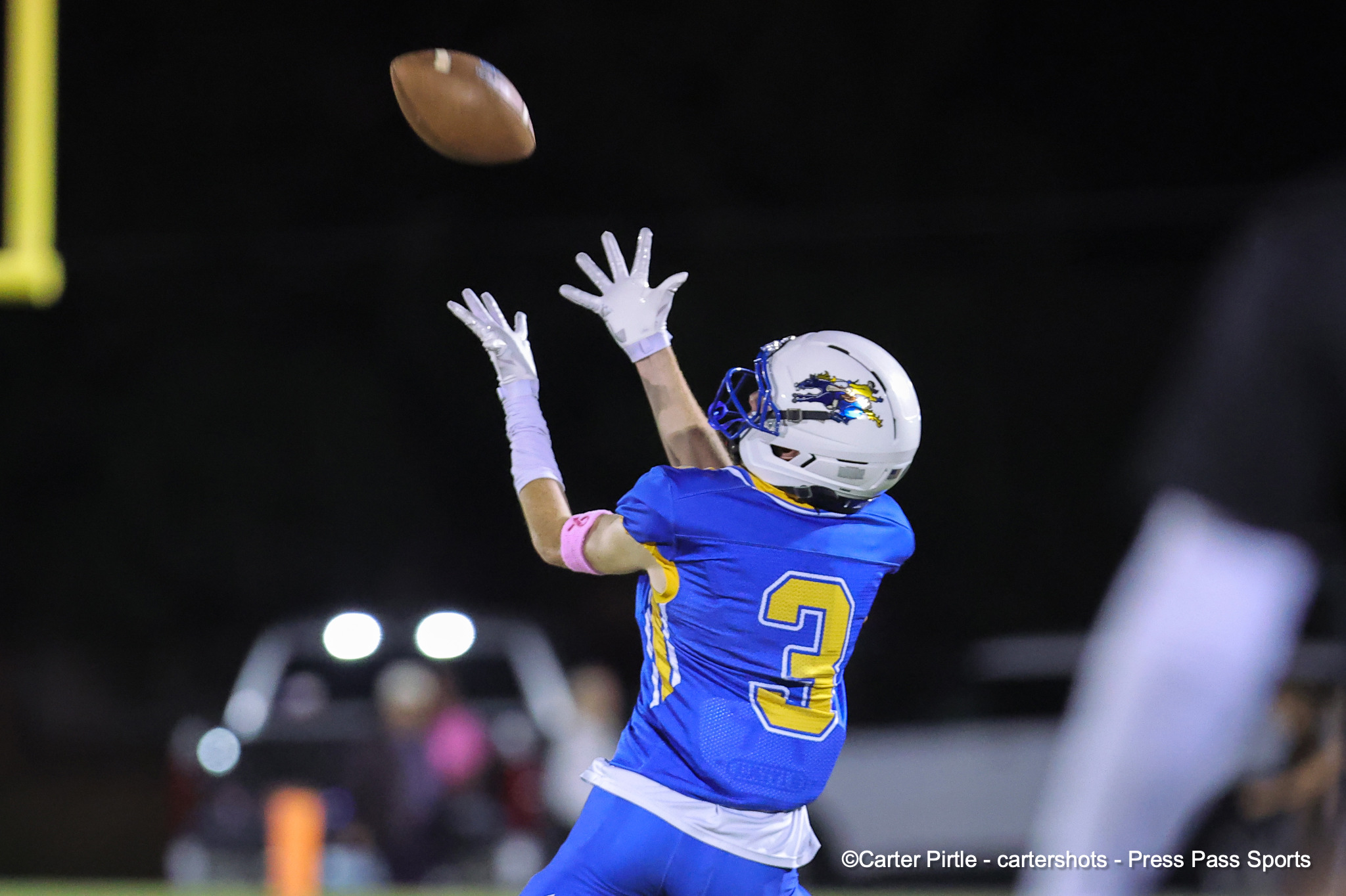 Football player in a blue uniform jumps to catch a pass at night, wearing white gloves and a helmet with a yellow-numbered jersey (number 3).