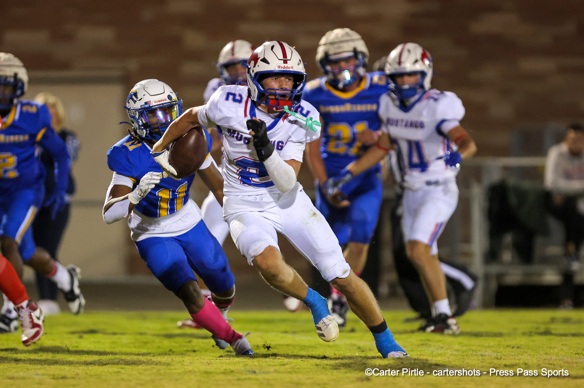 White-jersey football player holding the ball rushes forward as blue-team defenders chase him on a grass field under stadium lights
