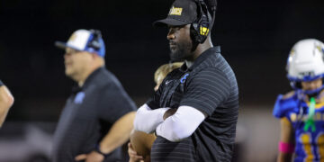 Coach on the sideline at night wearing a headset and a black cap, arms crossed.