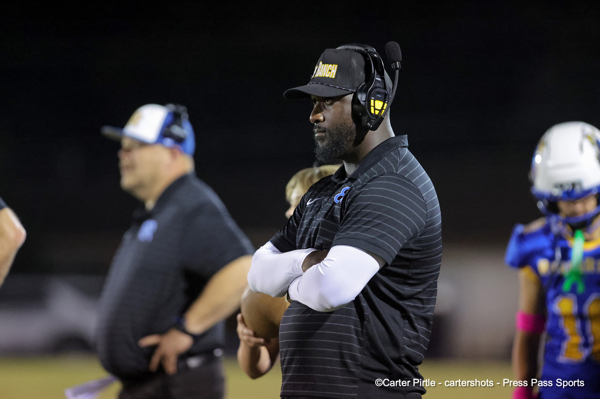 Coach on the sideline at night wearing a headset and a black cap, arms crossed.