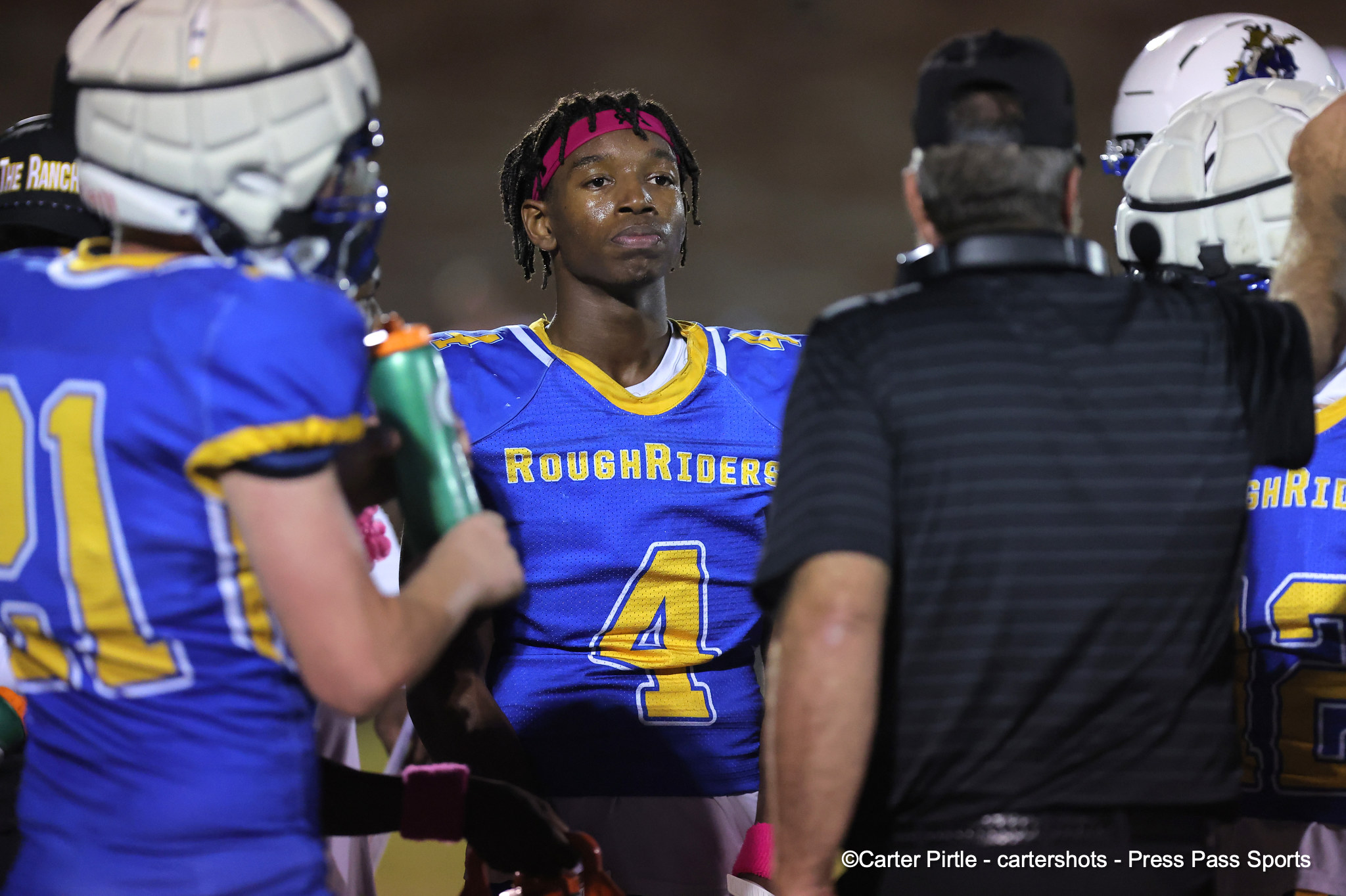 Football players in blue and gold RoughRiders uniforms huddle on the field at night, number 4 visible on the central player’s jersey.