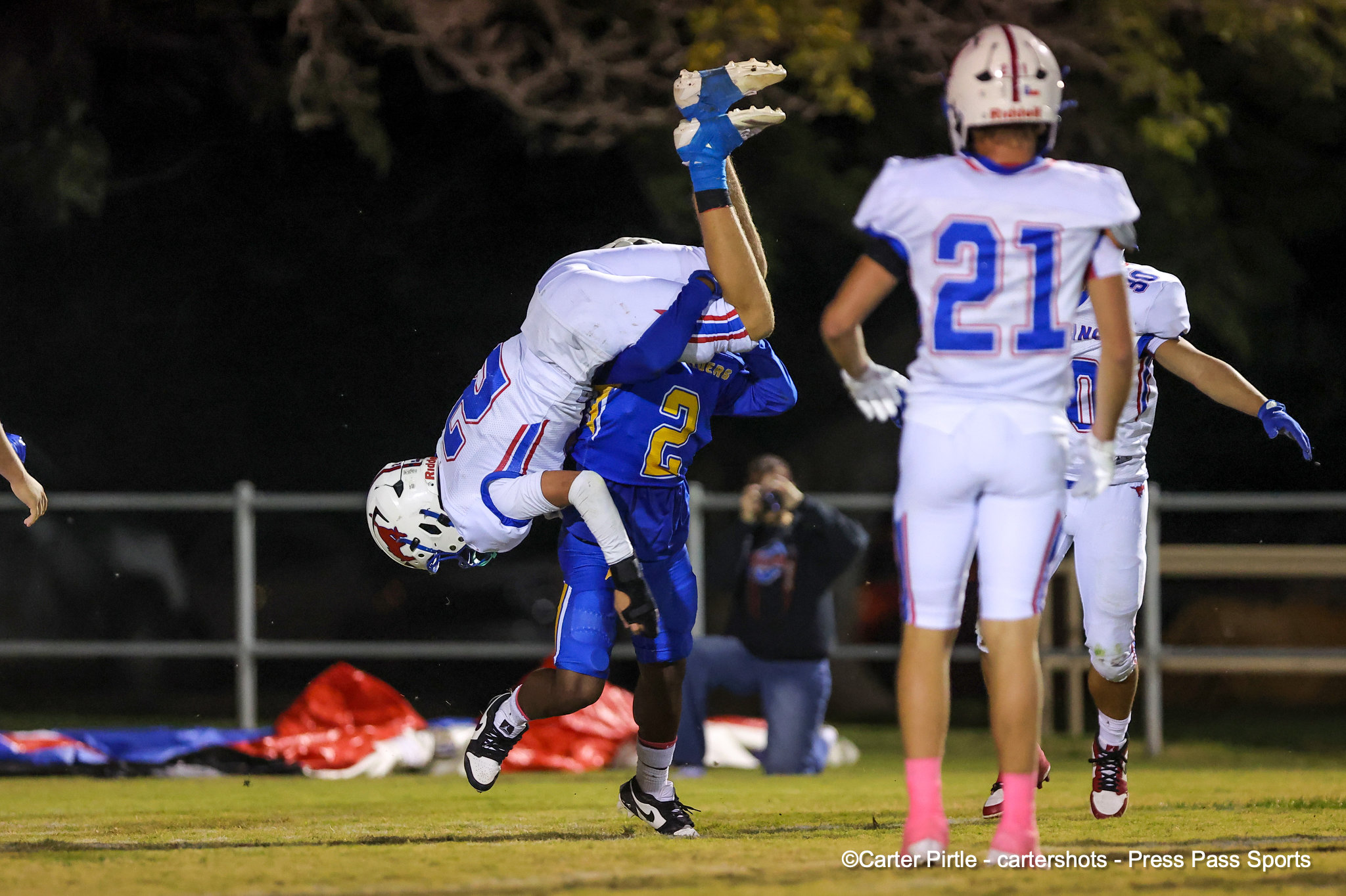 Nighttime football action as a blue-uniformed defender tackles a white-uniformed player who is mid-air, upside down near the ground on a grassy field.