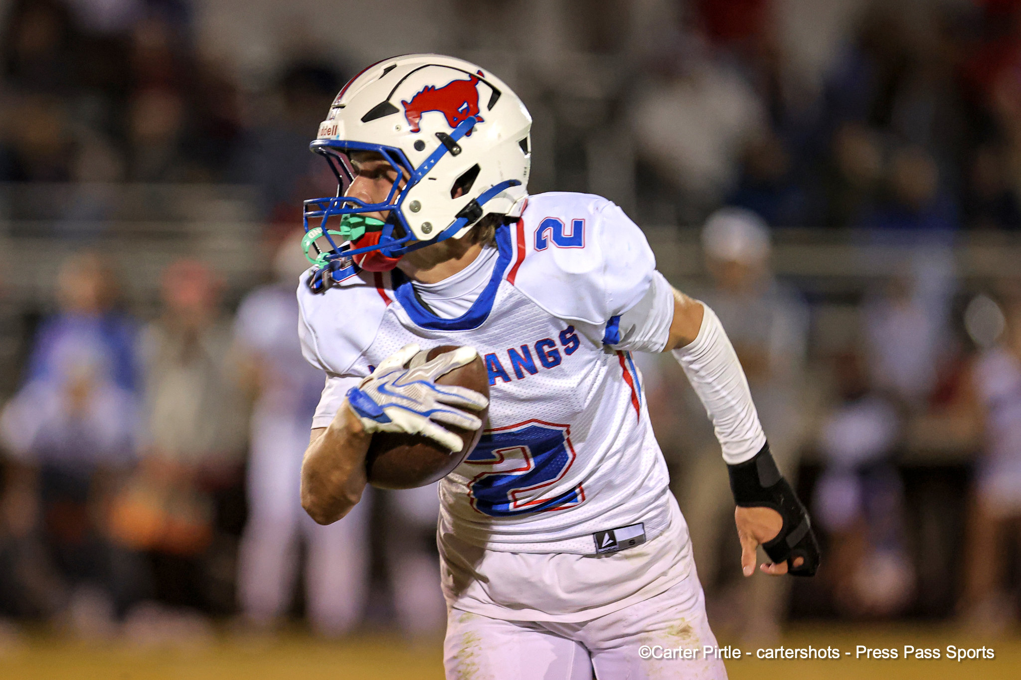Football player in a white uniform runs with the ball during a game, blue facemask and red logo helmet visible.