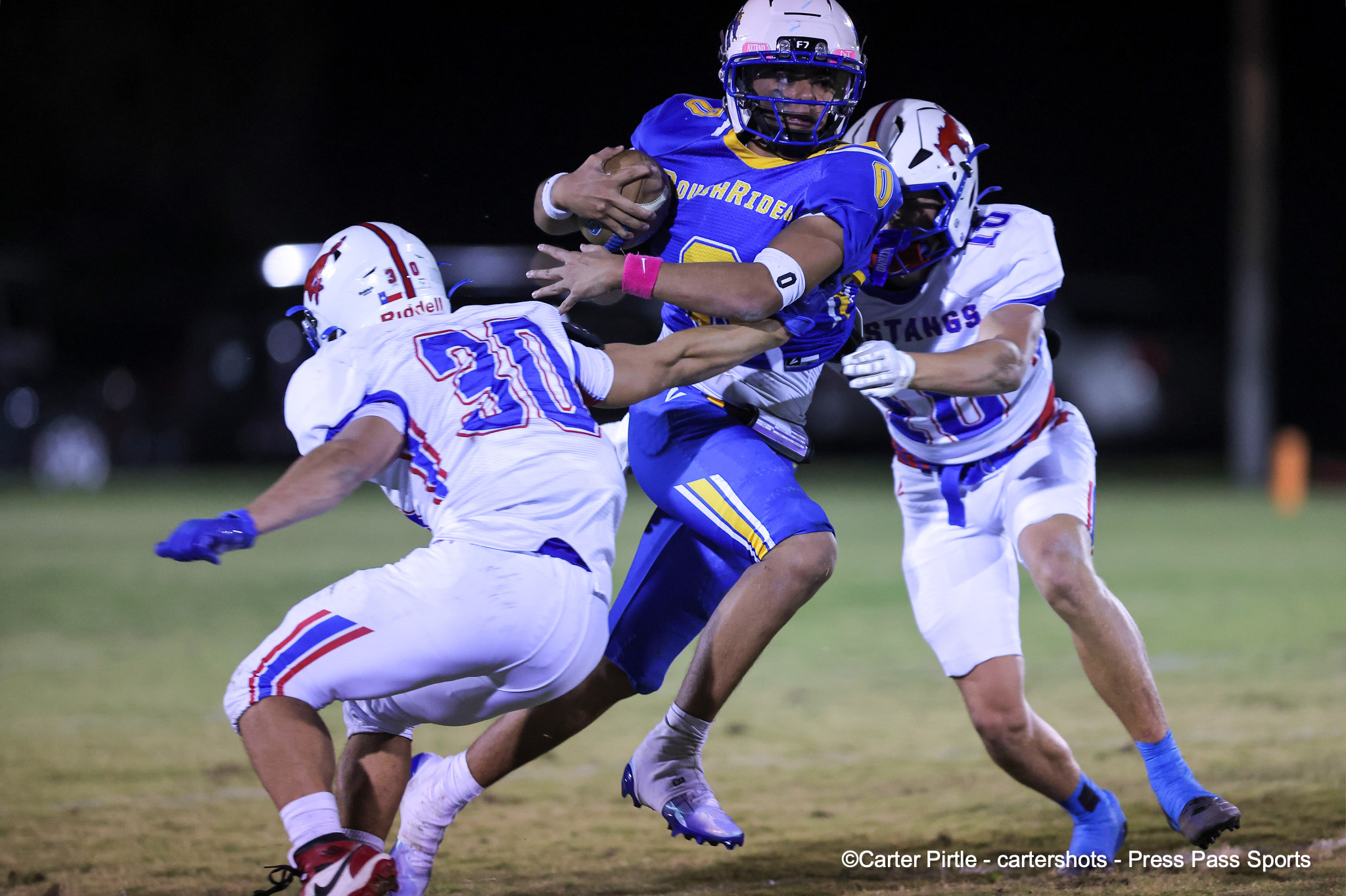 A football player in a blue uniform runs with the ball while two defenders in white uniforms tackle him during a night game.