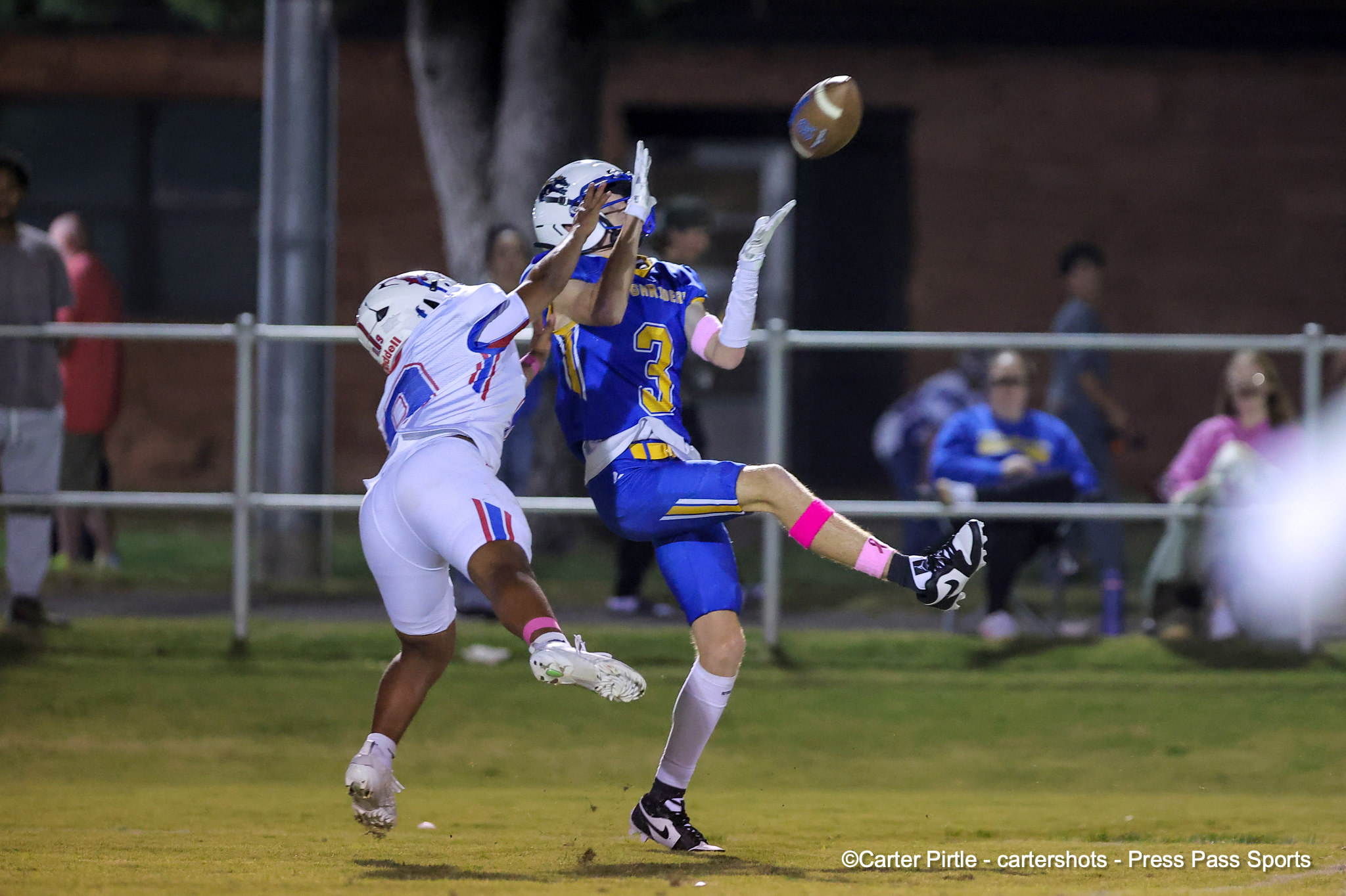 Two football players leap for the ball during a night game, one in blue and yellow and the other in white, near the sideline.