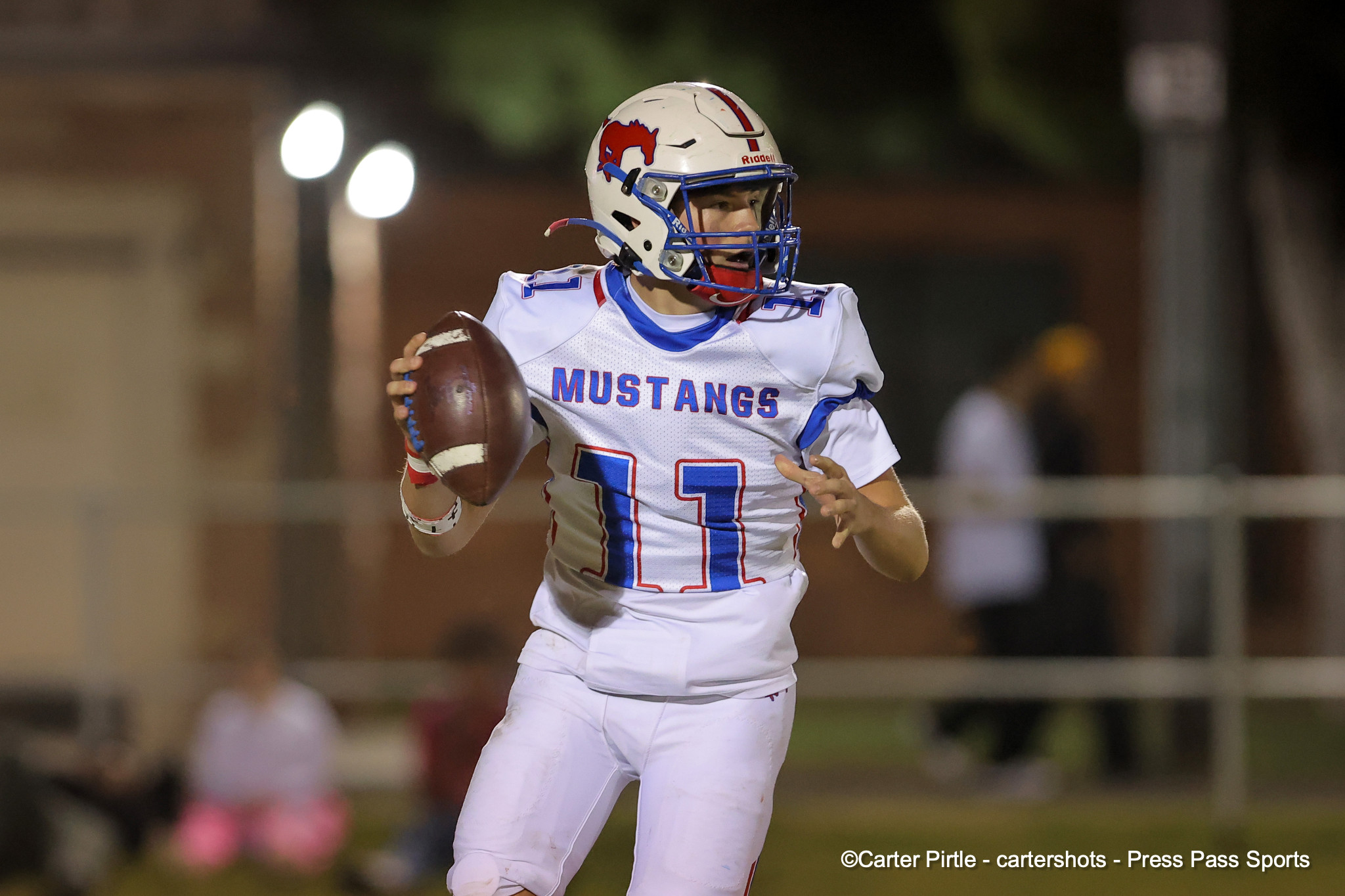 Football player in a white blue-and-red Mustangs uniform runs with the ball on a nighttime field.