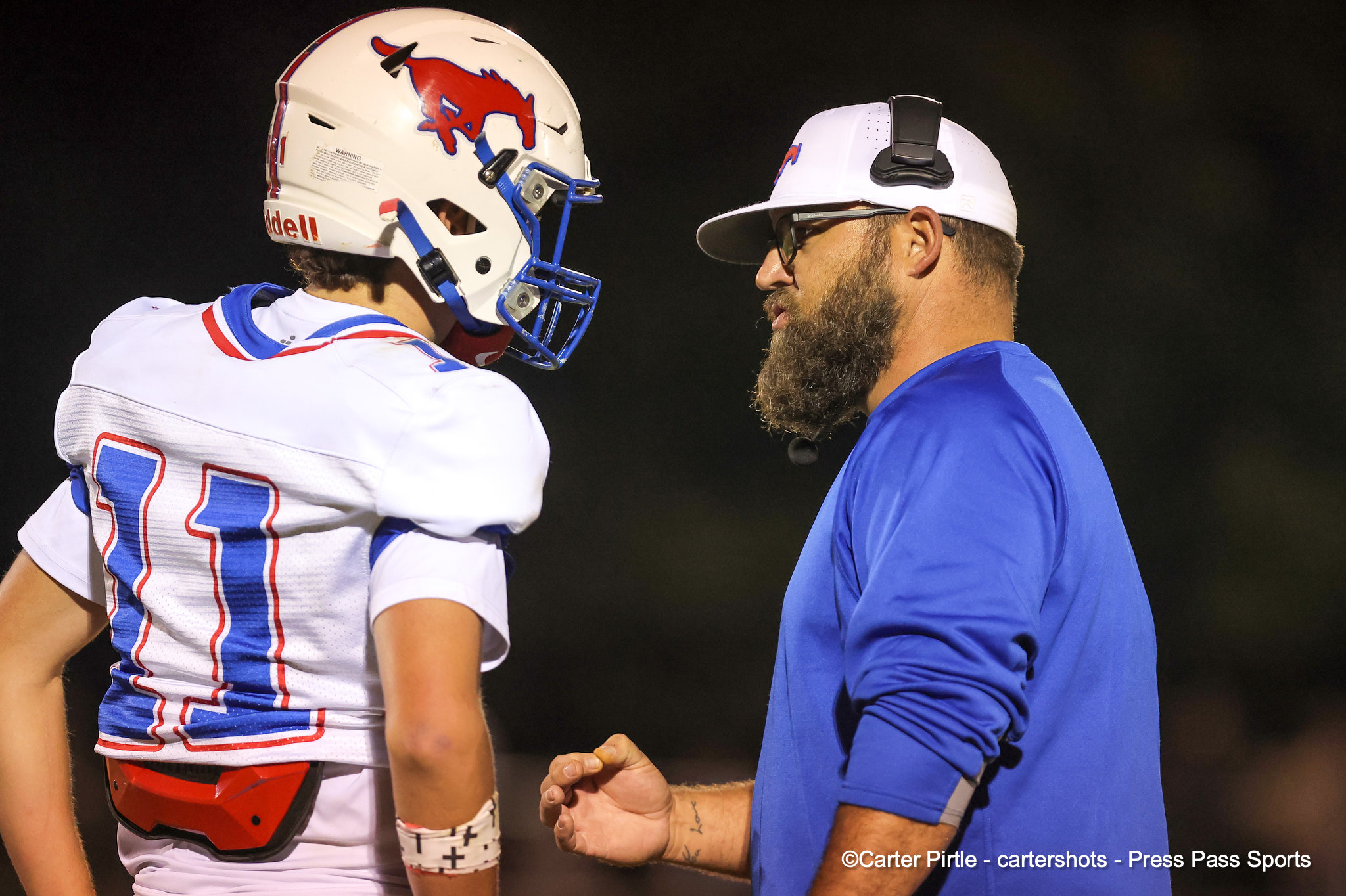 Football player in a white jersey and helmet facing a bearded coach in a blue shirt and white cap, about to fist bump on a dark field.