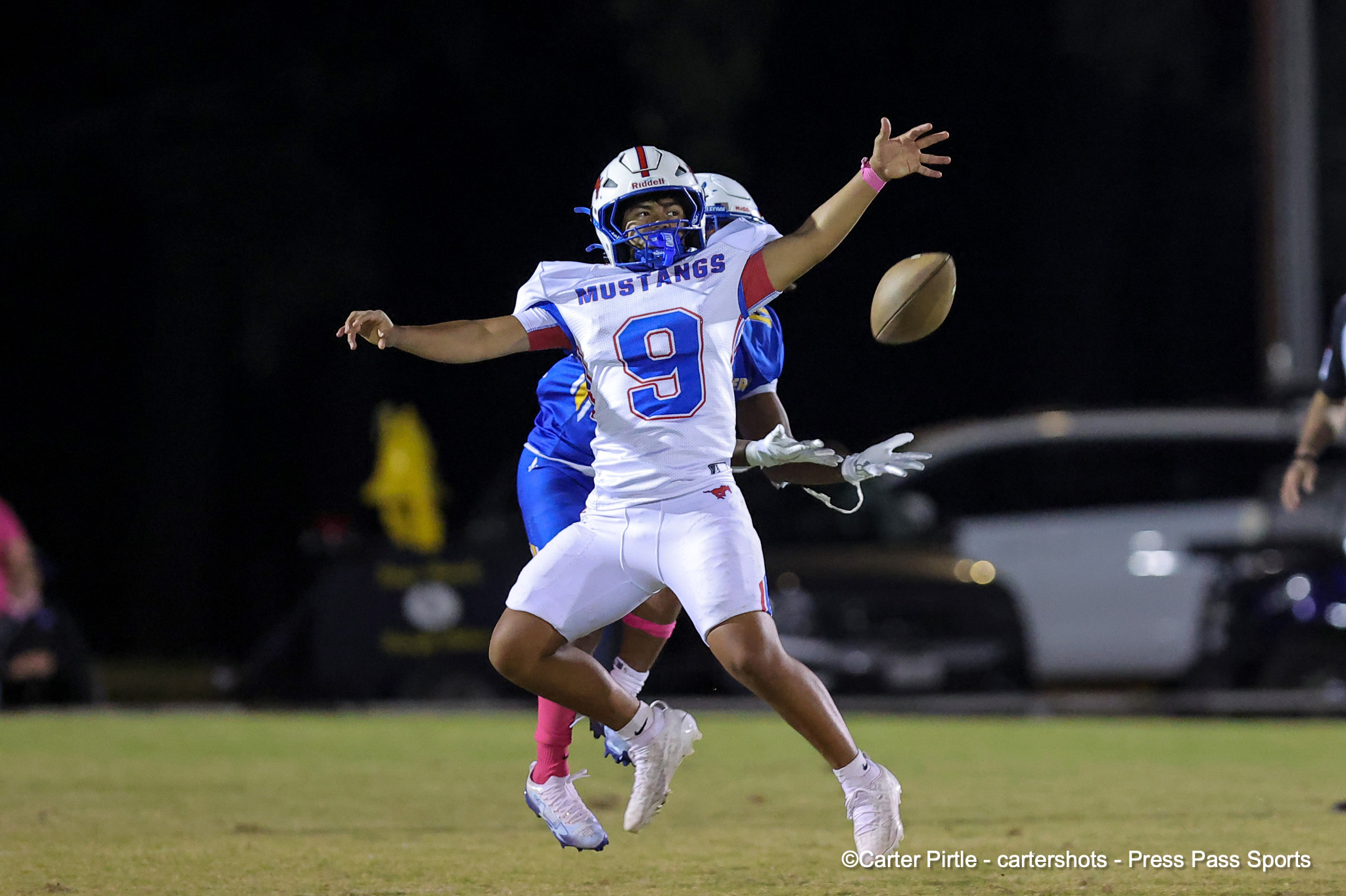 Two football players leap for a ball during a night game; white jersey with'MUSTANGS' and number 9 is visible in foreground.
