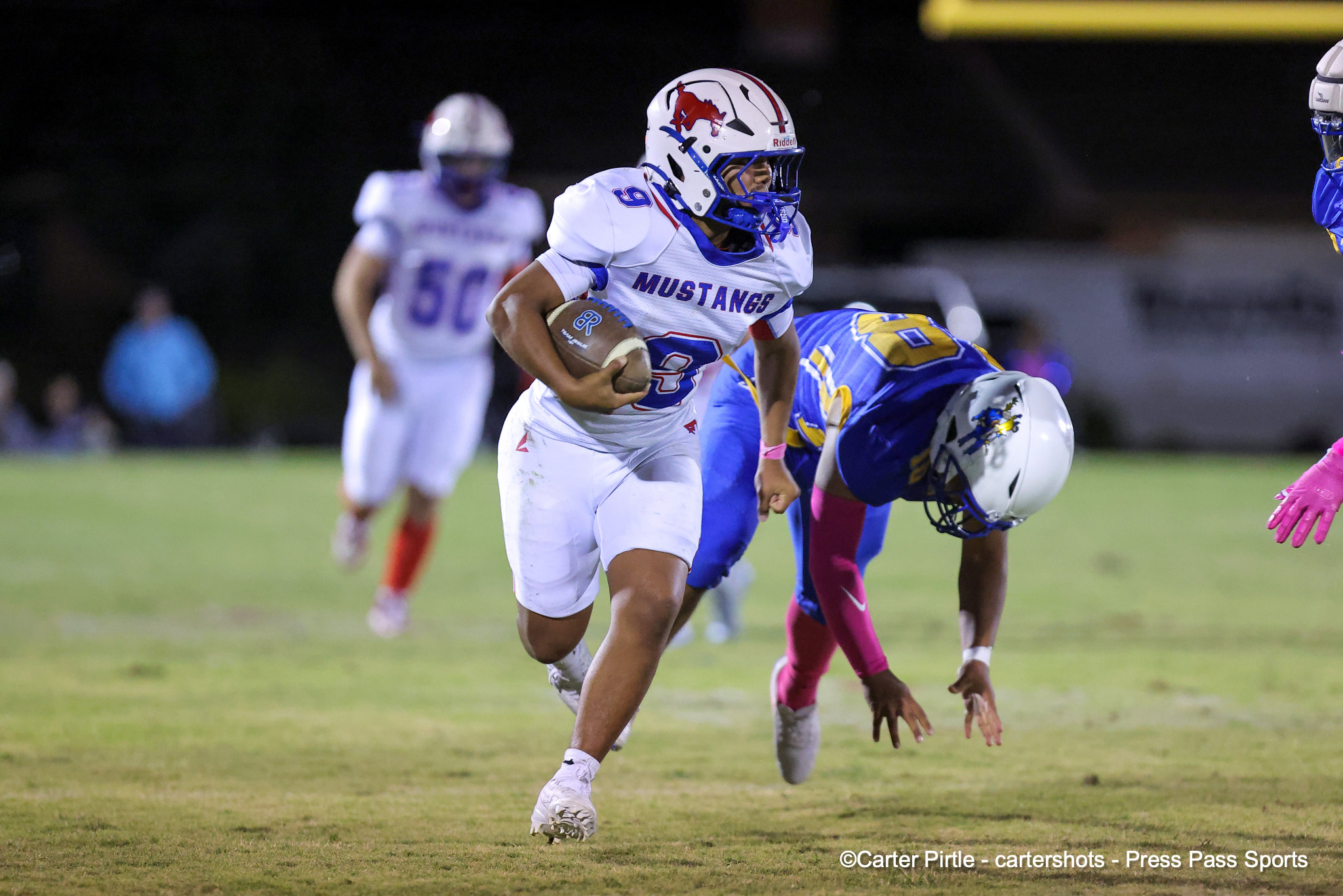 Football game action: a player in a white Mustang uniform runs with the ball at night on a grass field, while opponents pursue.
