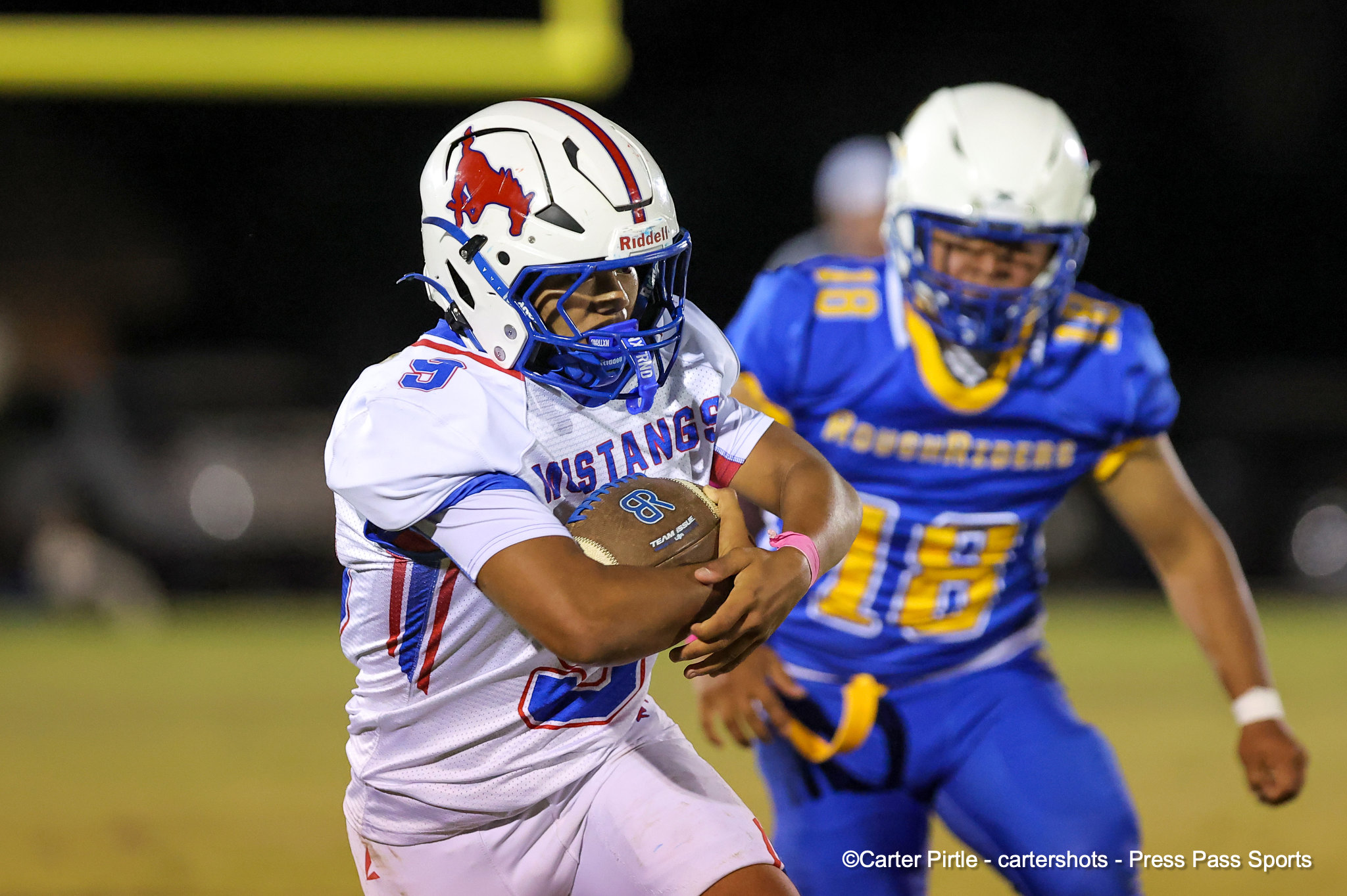 A football running back in a white uniform and helmet with a red Mustang logo carries the ball while a blue-and-yellow defender chases behind at night.