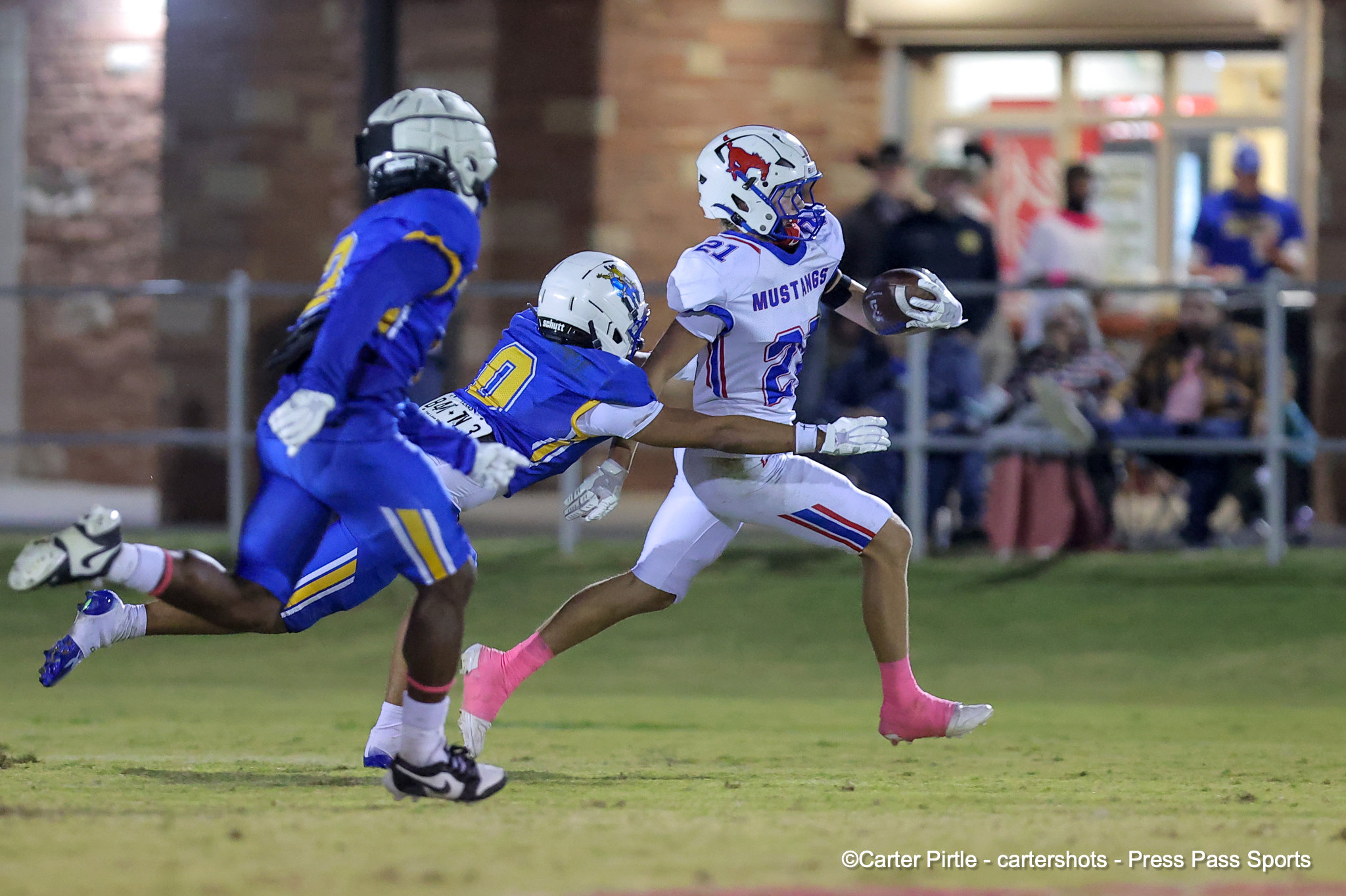 Lacrosse player in white Mustang jersey runs with the ball as a blue-clad defender pursues on a grass field, with a blurred crowd in the background.