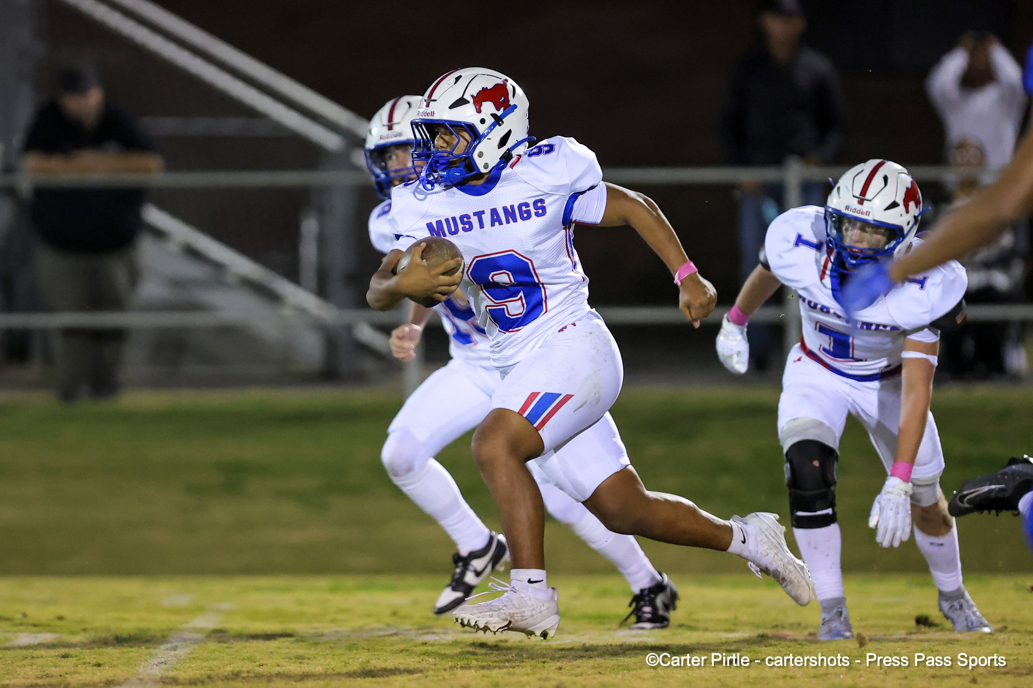 Football action: a Mustang runner with the ball sprinting forward while teammates chase during a night game on a grass field.