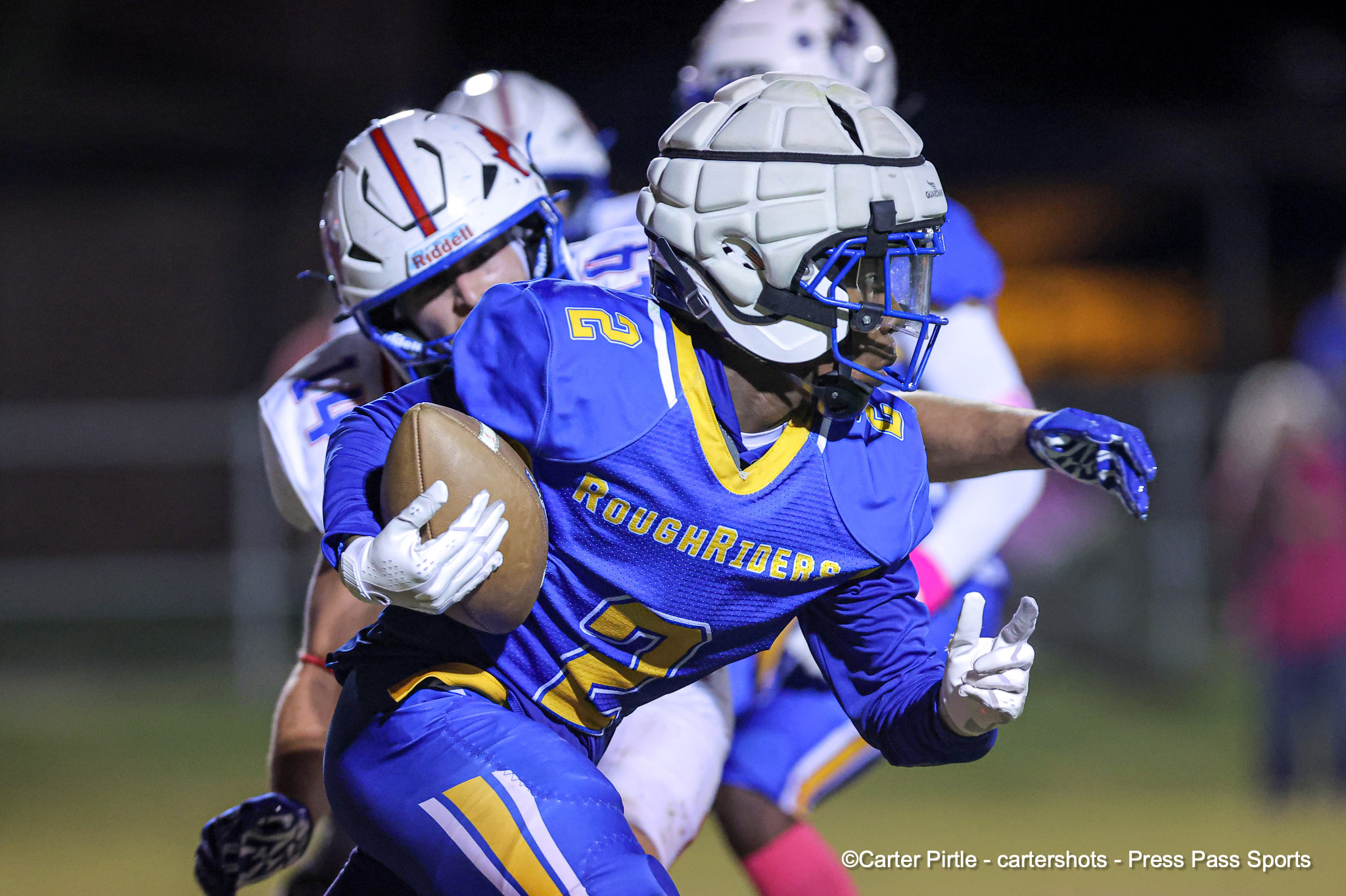 Blue-clad football players chase on a night game; a ball carrier in front holds the ball tightly as teammates pursue.