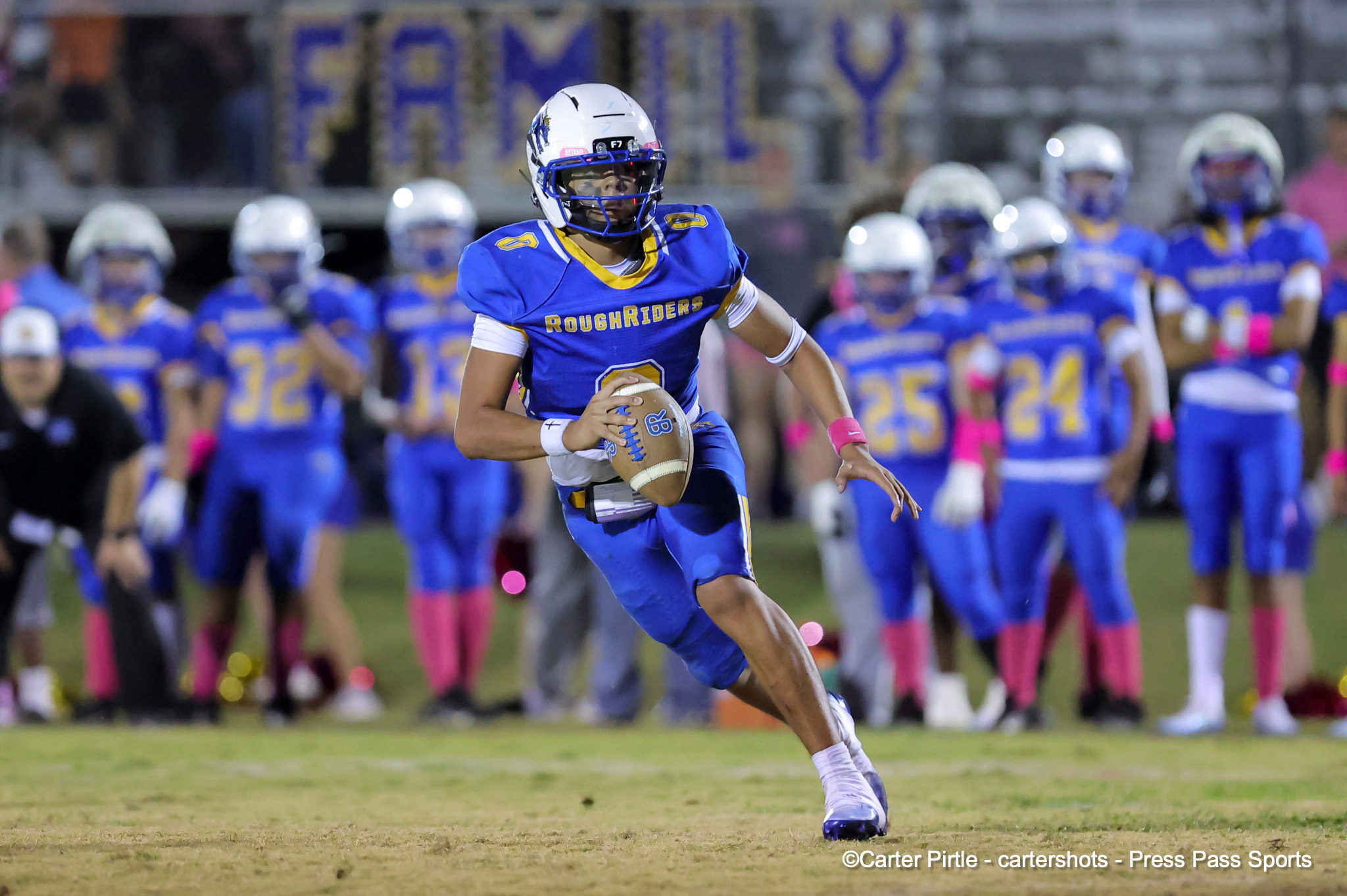 RoughRiders player in blue and gold uniform runs with the football toward the camera as teammates look on in the blurred background.