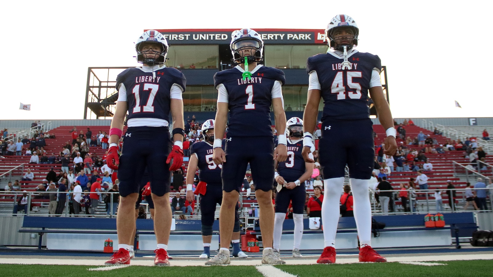 Three Liberty football players stand on the field before a game, wearing navy uniforms numbered 17, 1 and 45 with a stadium and crowd behind them.