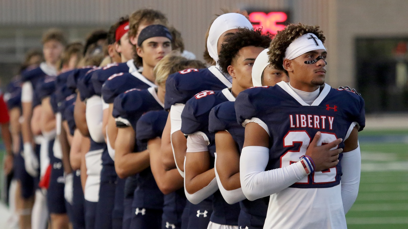 Line of high school football players in navy uniforms with white accents, arms linked in a pre-game or ceremonial line on a green turf field.