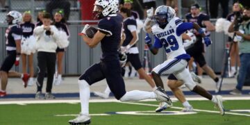 Navy-clad football player with the ball runs toward the end zone as a blue-and-white defender chases him.
