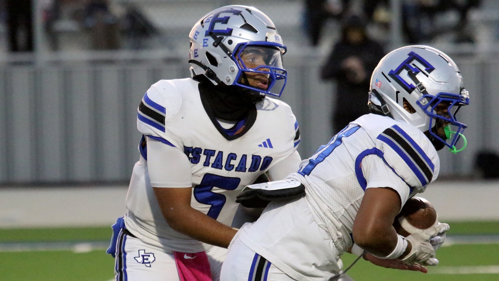 Two football players in white jerseys with blue trim tackle on a green field; one guards the ball carrier.