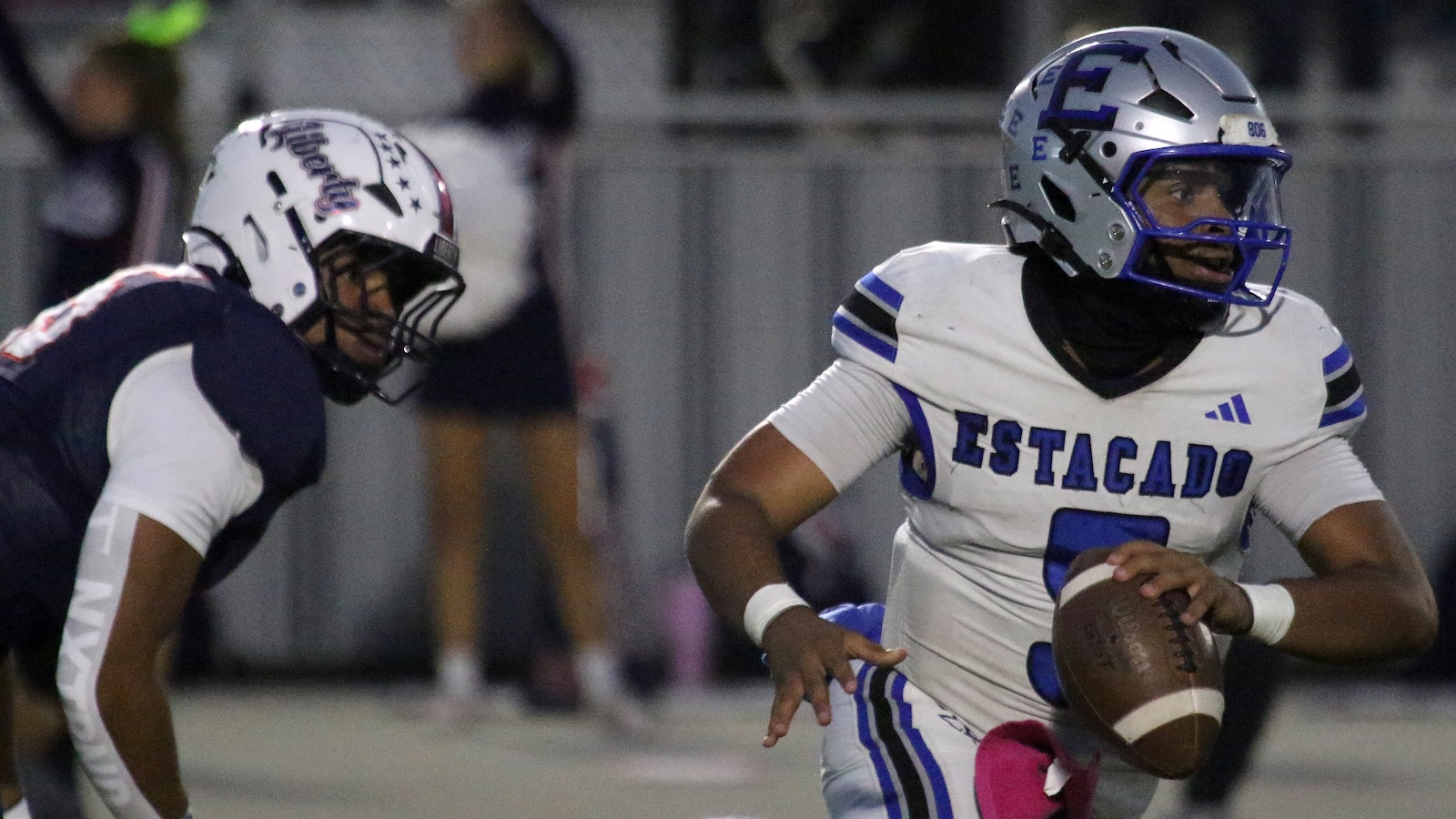 Football player in white and blue with'ESTCADO' (likely ESTACADO) holding a football and running as a defender approaches.