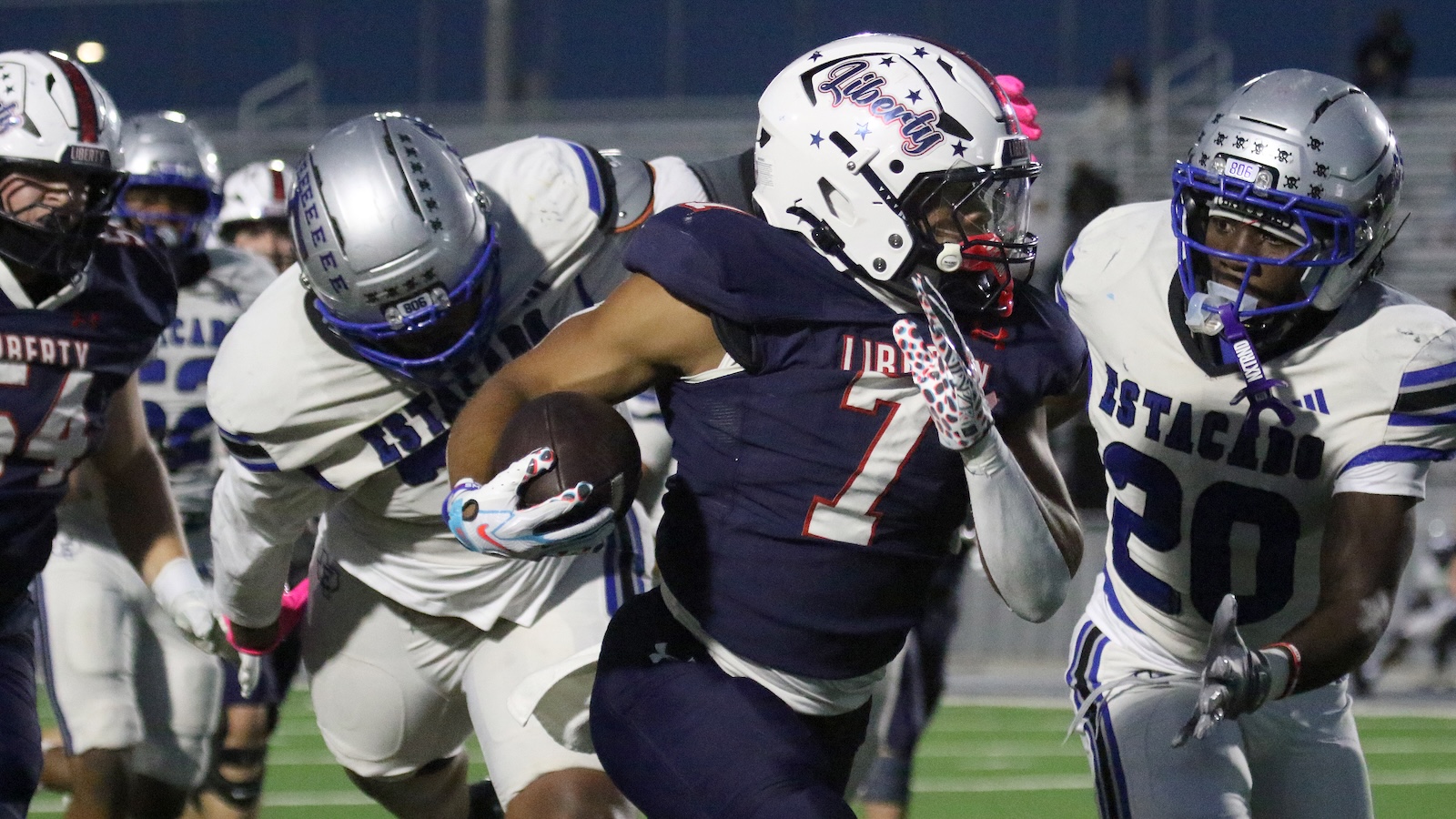 Navy-clad football player wearing number 7 carries the ball as defenders chase during a night game.