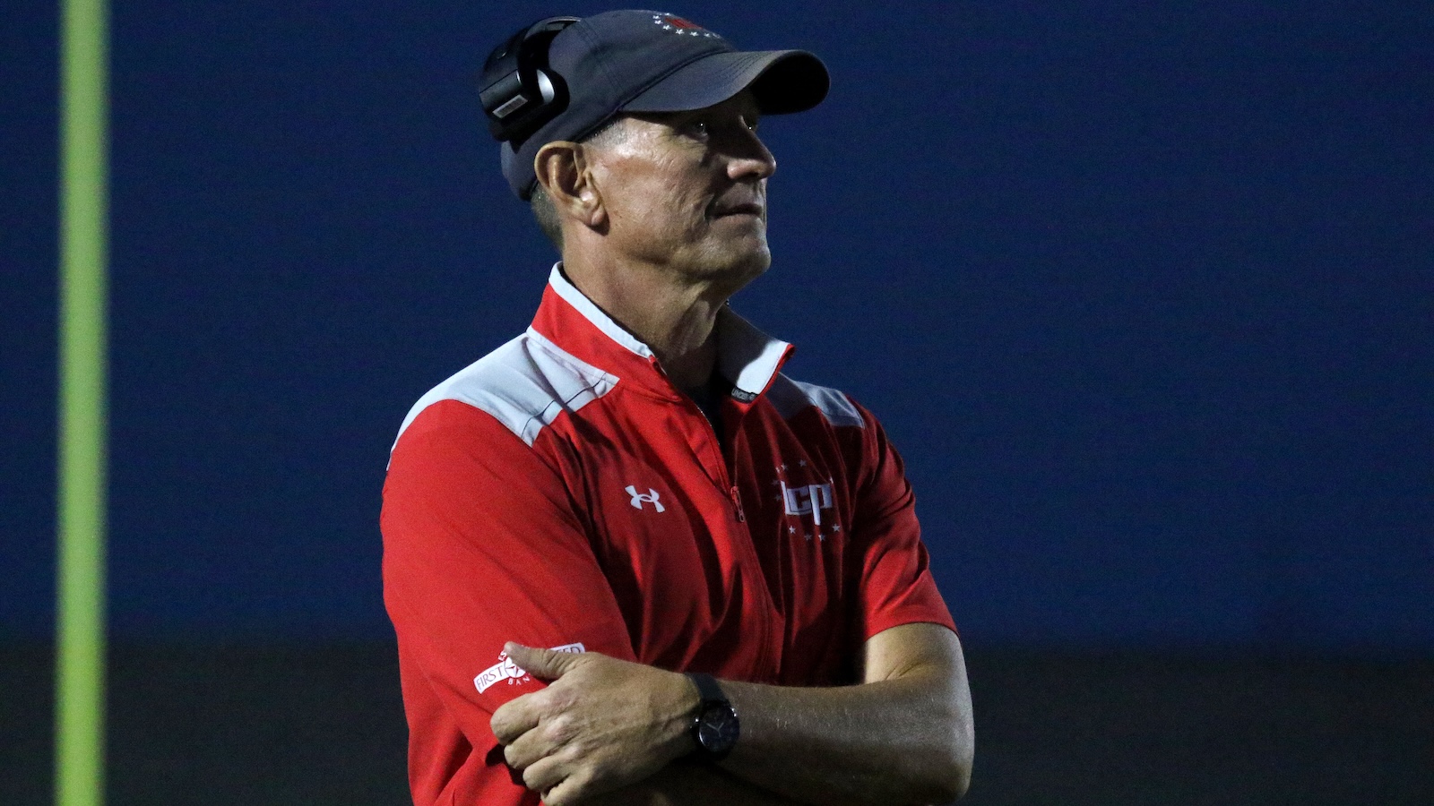 Coach standing with arms crossed on a dark sports field at night, wearing a red Under Armour jacket, a cap, and headset.
