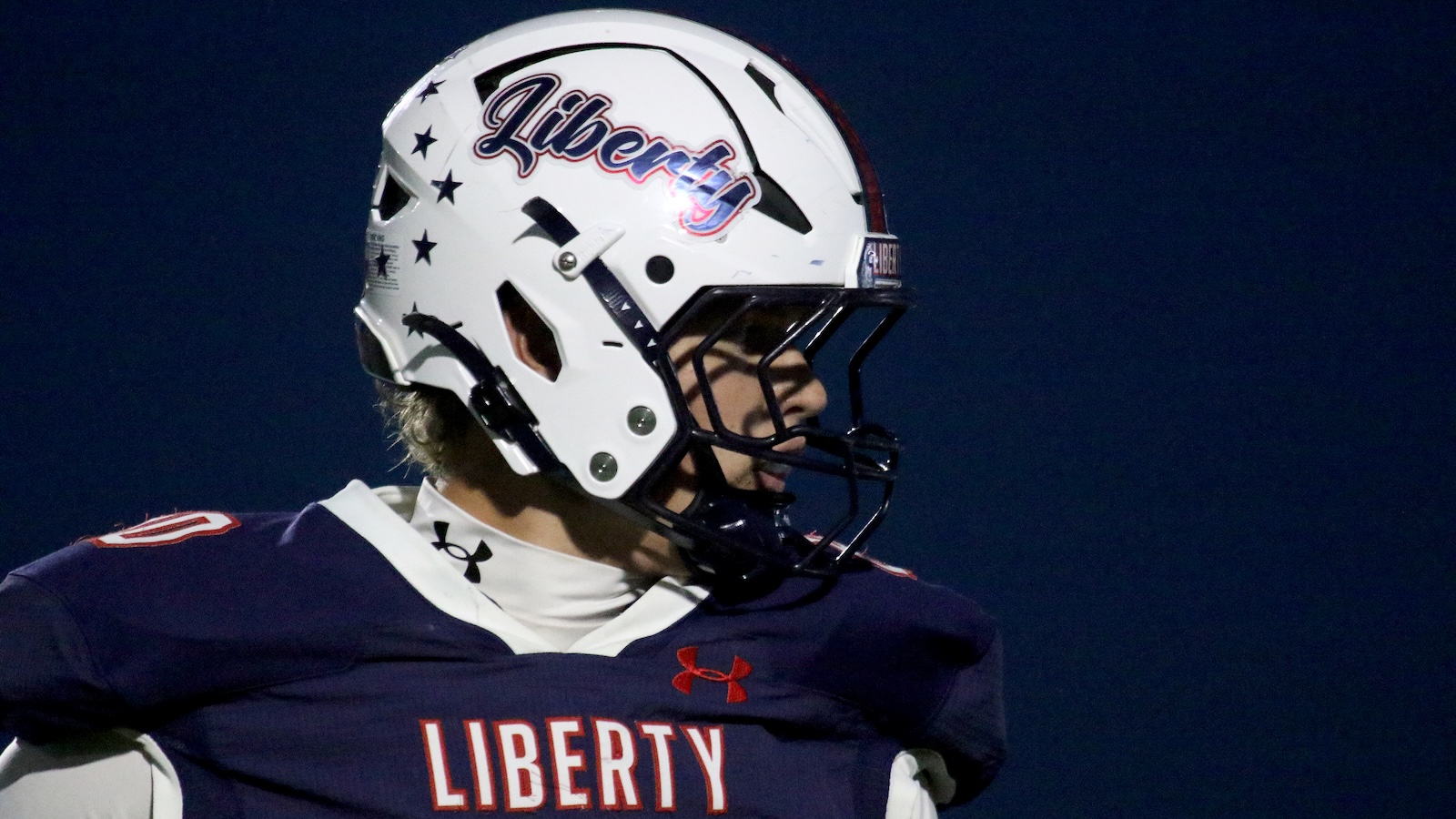 Close-up of a football player in a Liberty uniform, wearing a white helmet with'Liberty' script and star decals, looking to the right.