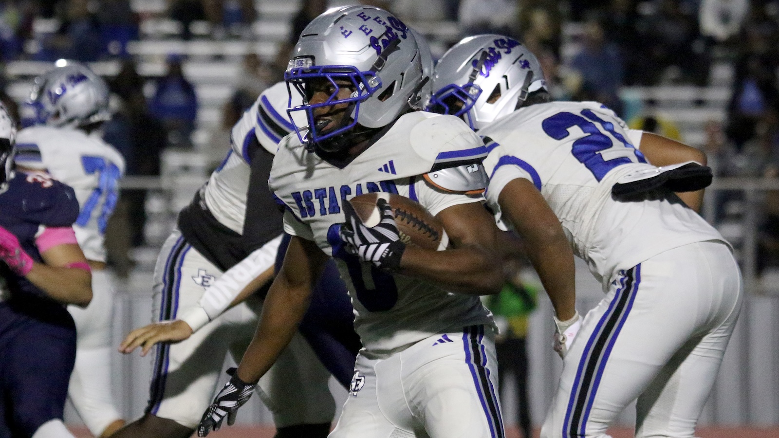 Football player in white and blue uniform runs with the ball during a night game, tackled by teammates and opponents nearby, silver helmet with blue faceguard visible