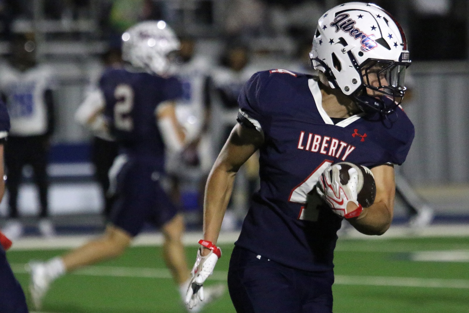 Liberty football player in a navy jersey runs with the ball on a green field at night, white helmet with Liberty logo visible.