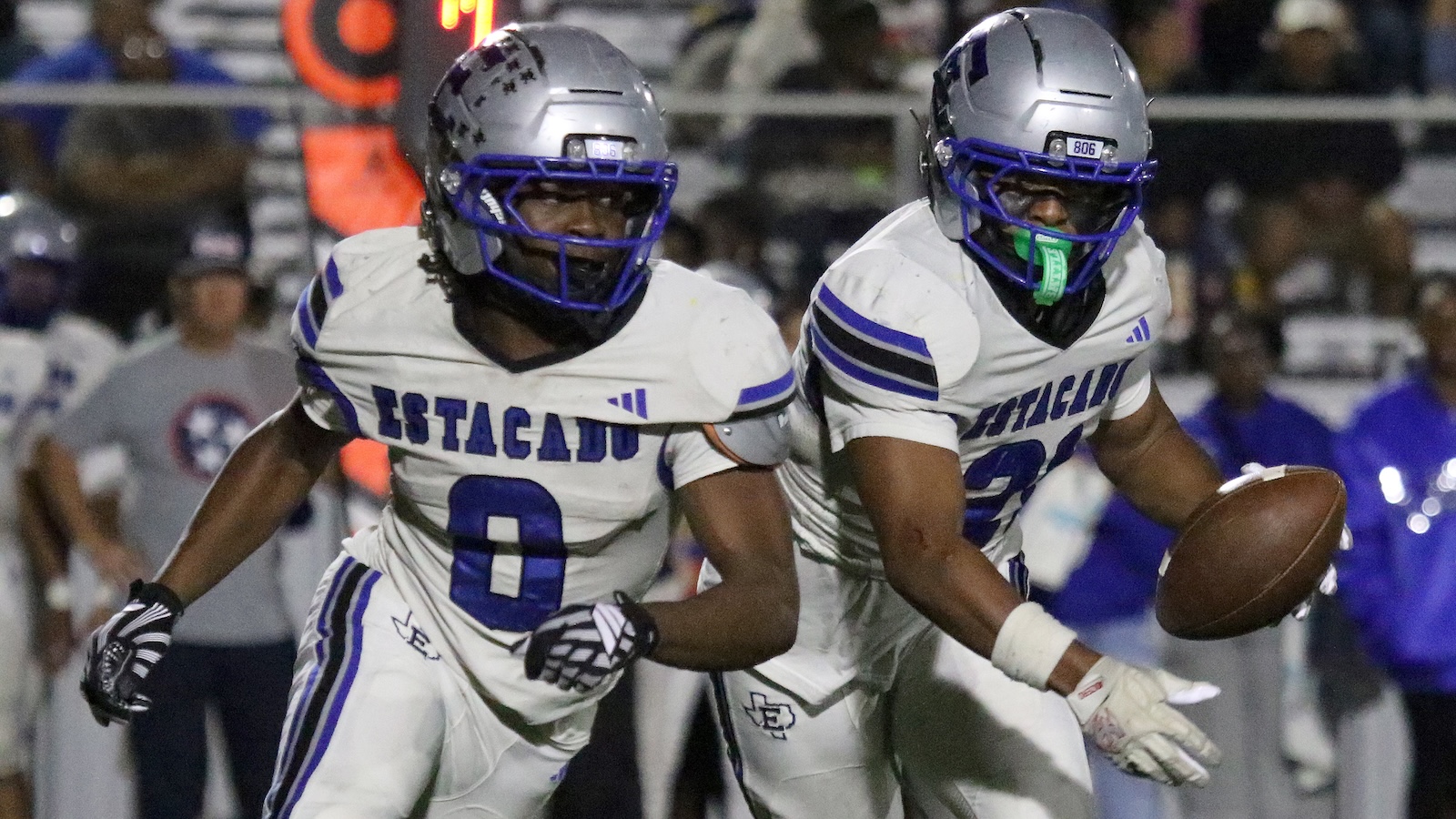 Two football players in white and blue uniforms run on the field during a night game, one carrying the ball.
