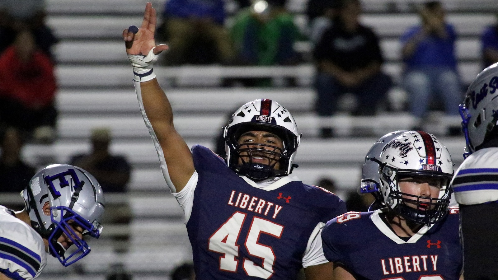 Football player from Liberty raises his right arm in triumph during a game at night. The navy jersey shows number 45 and a white helmet with stars.