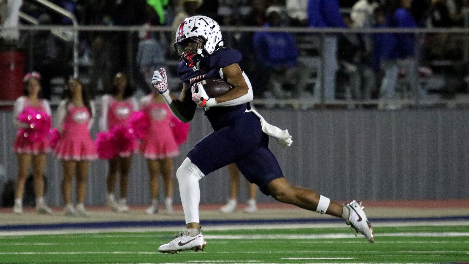 Football player in navy uniform running with the ball on a well-lit field; pink-clad cheerleaders line the sideline in background.