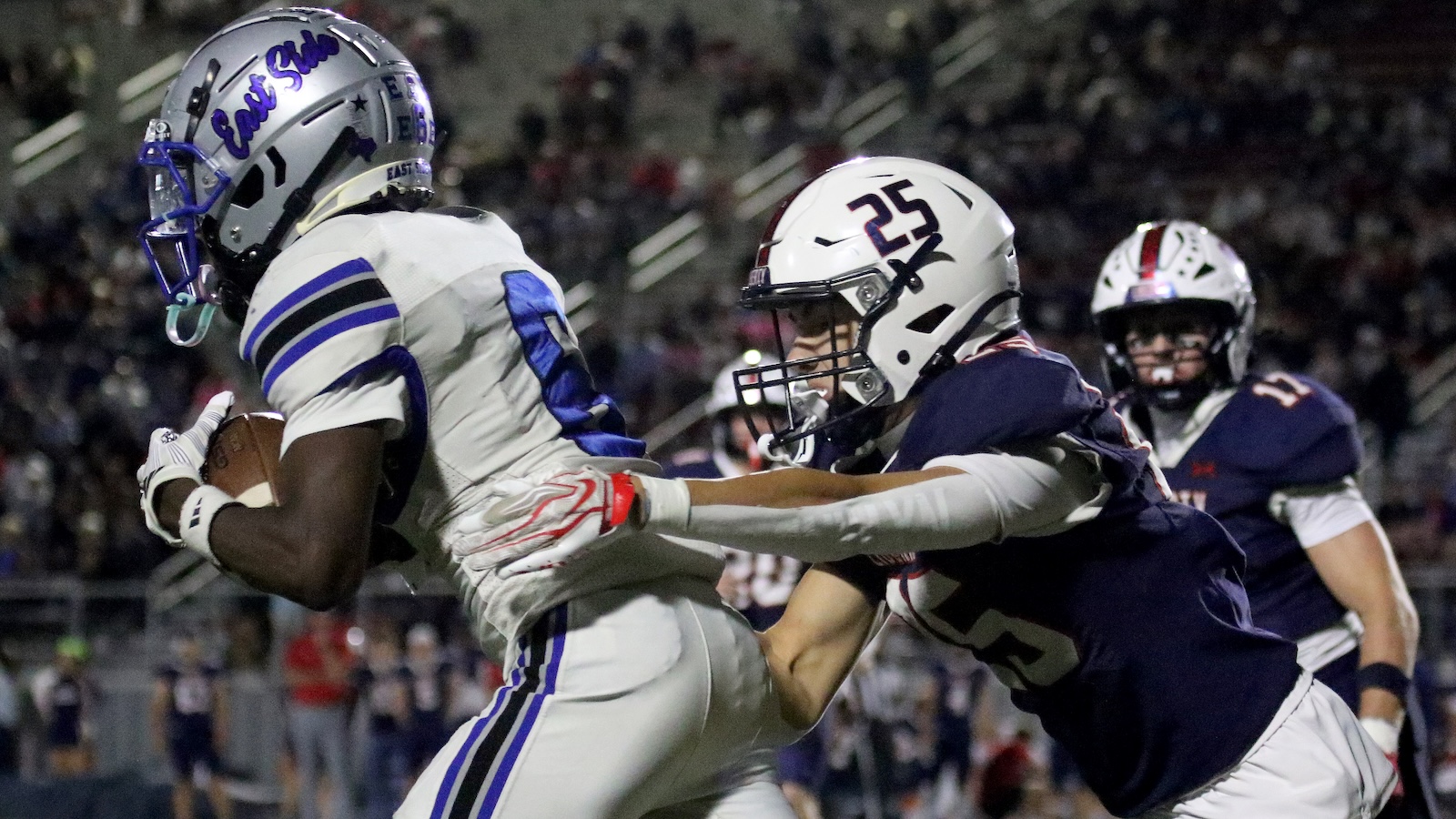 Football action: a ball carrier in white and blue advances with the ball while a defender in navy reaches to tackle him.