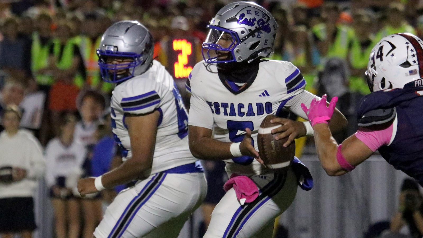 Quarterback in a white Estacado jersey holding the ball, looking for a play as a defender in pink gloves reaches in during a night game.