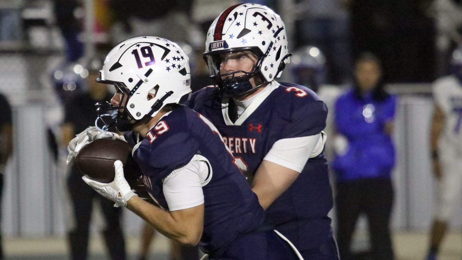Two Liberty University football players in navy uniforms tackle near the sideline, with one grabbing the football.
