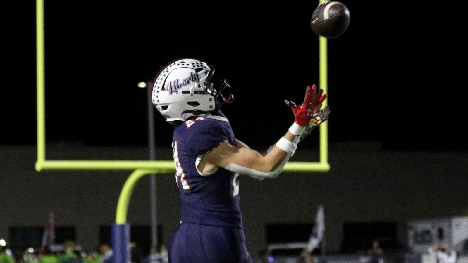 Football player in navy uniform and white star-pattern helmet extends arms to catch a pass near a bright yellow goalpost at night stadium light.