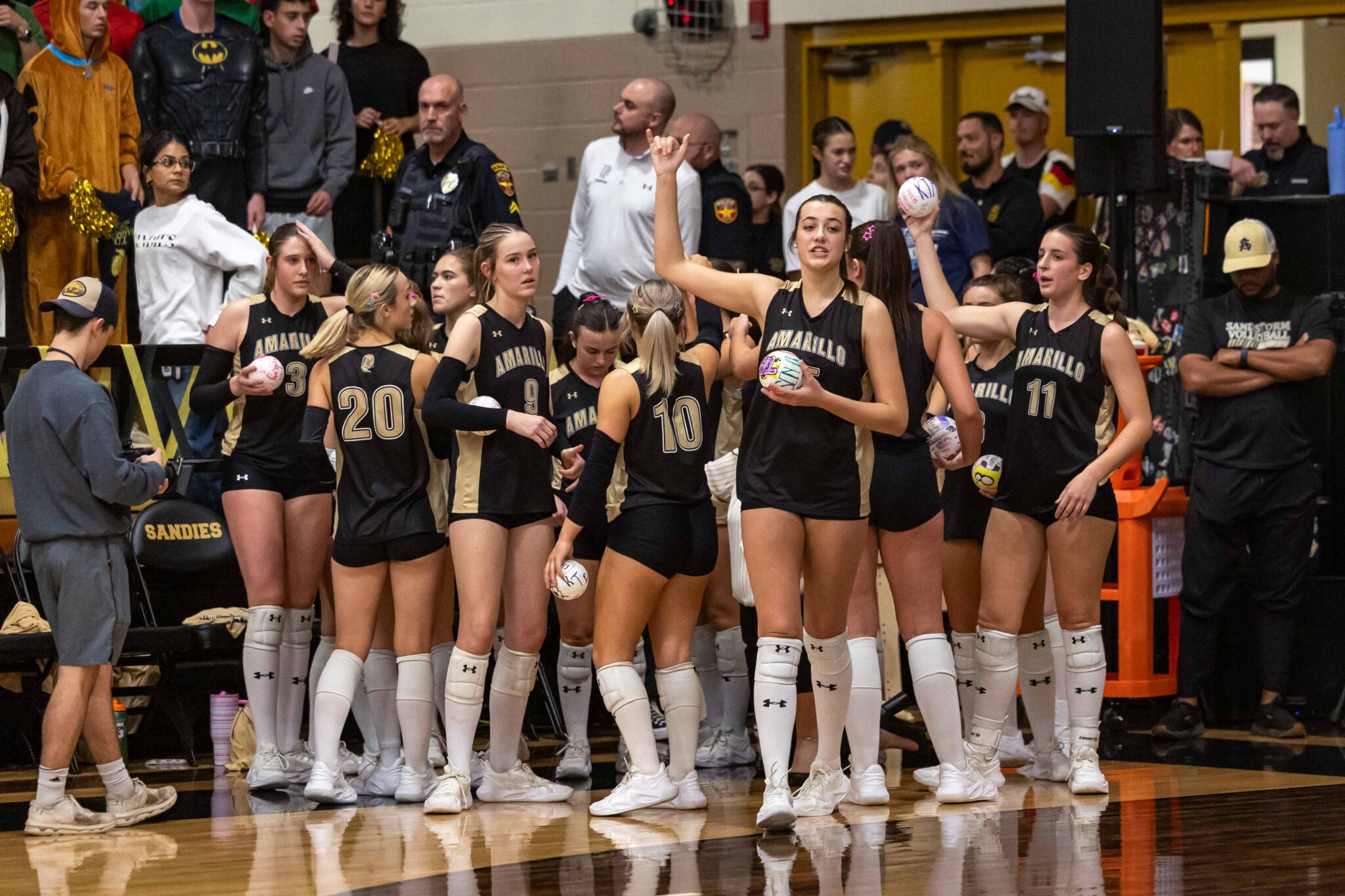 THS-AHS-VB-2 | Press Pass Sports Group of female volleyball players in black and gold uniforms standing together on a gym court, some holding volleyballs, with spectators in the background.