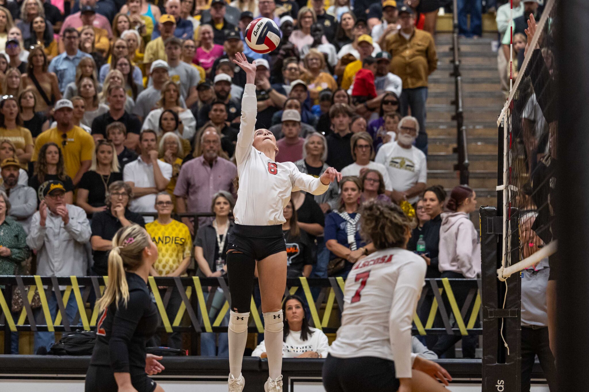 THS-AHS-VB-5 | Press Pass Sports Female volleyball player in a white uniform jumps to spike the ball as a crowded gym audience watches.