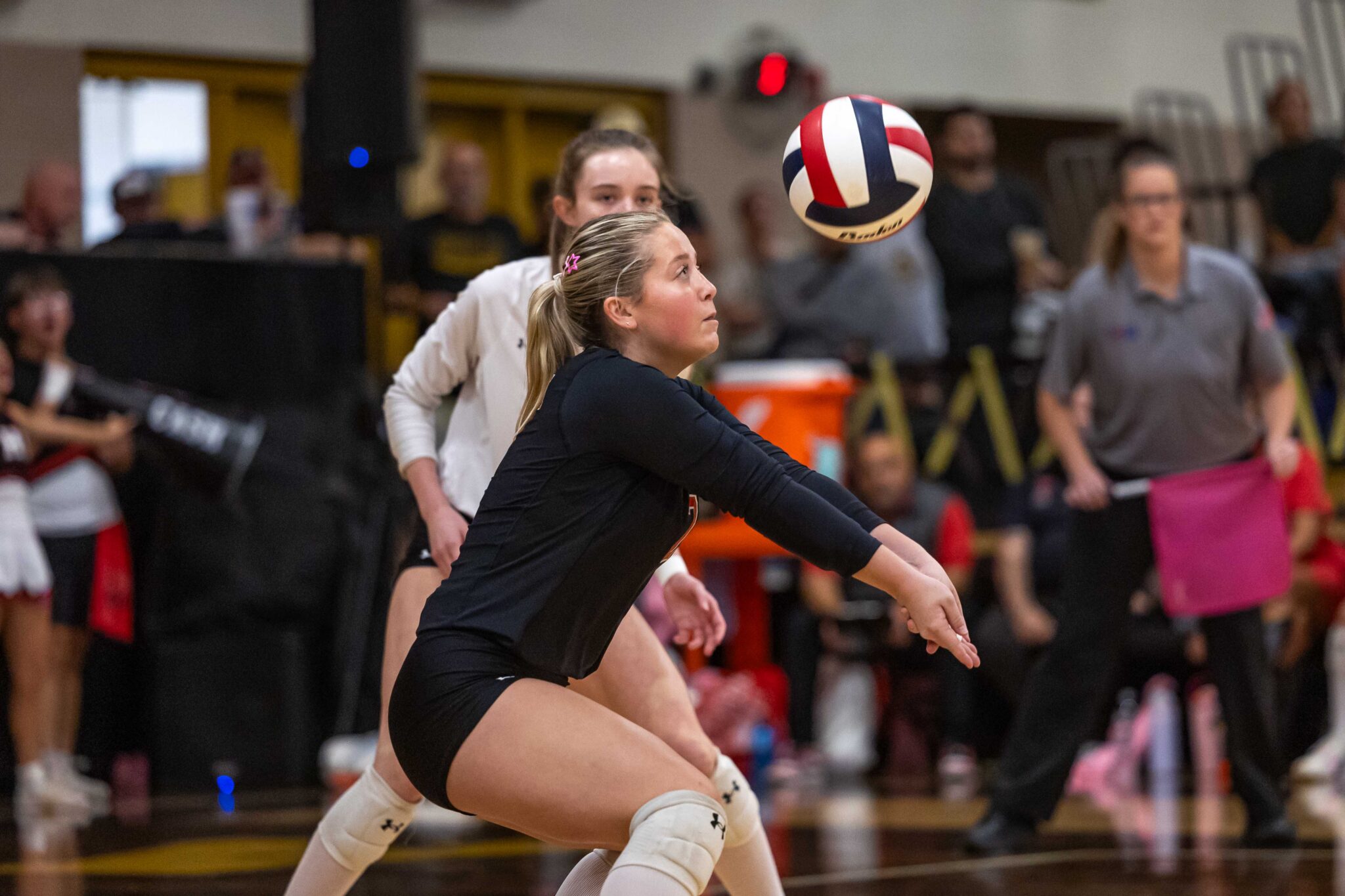 THS-AHS-VB-9 | Press Pass Sports Female volleyball players reach for a volleyball mid‑air during a match in a gym, with spectators in the background.