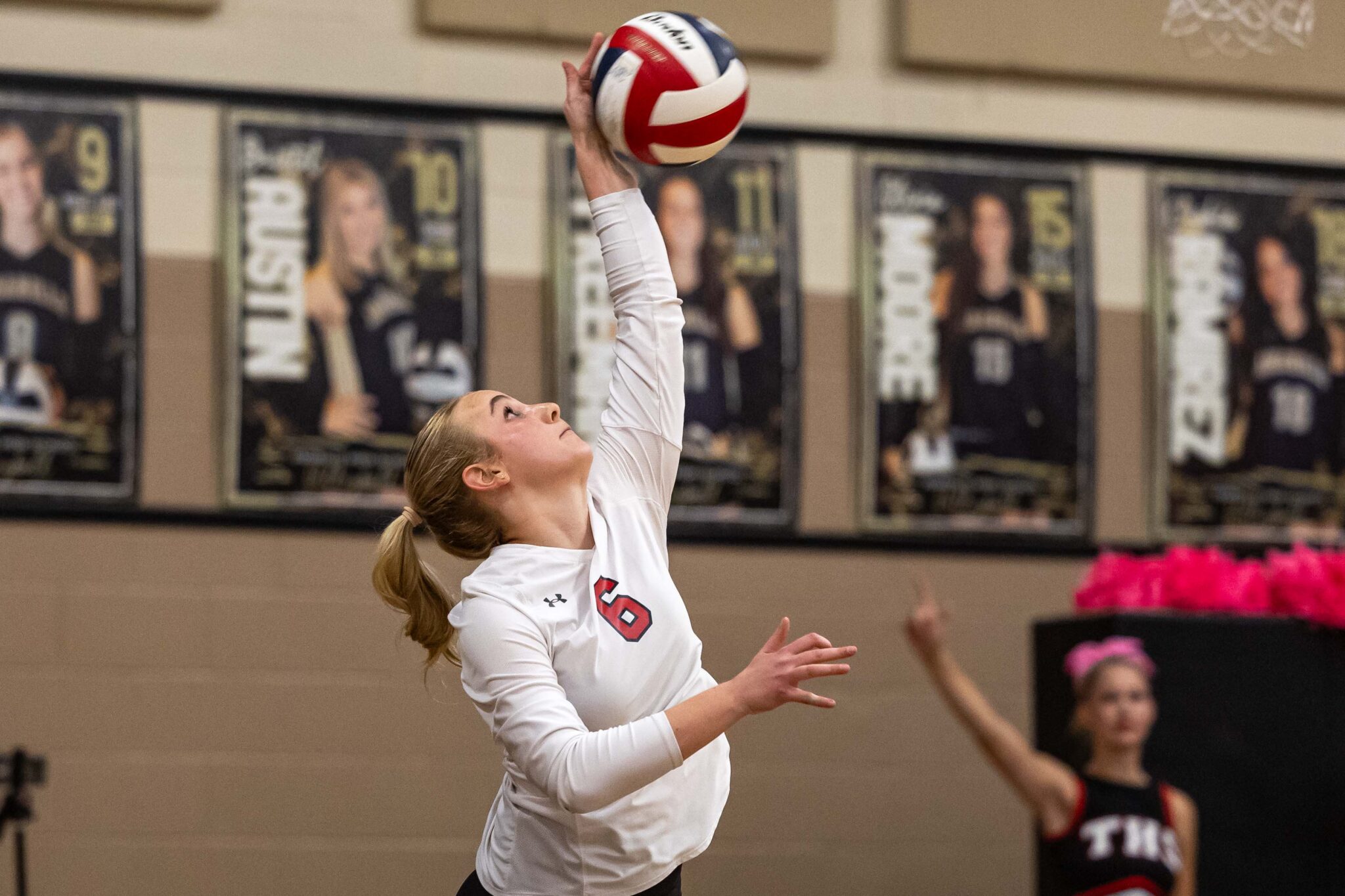 THS-AHS-VB-12 | Press Pass Sports Female volleyball player in a white jersey reaches up to hit a red‑white ball in an indoor gym, with banners/posters in the background.
