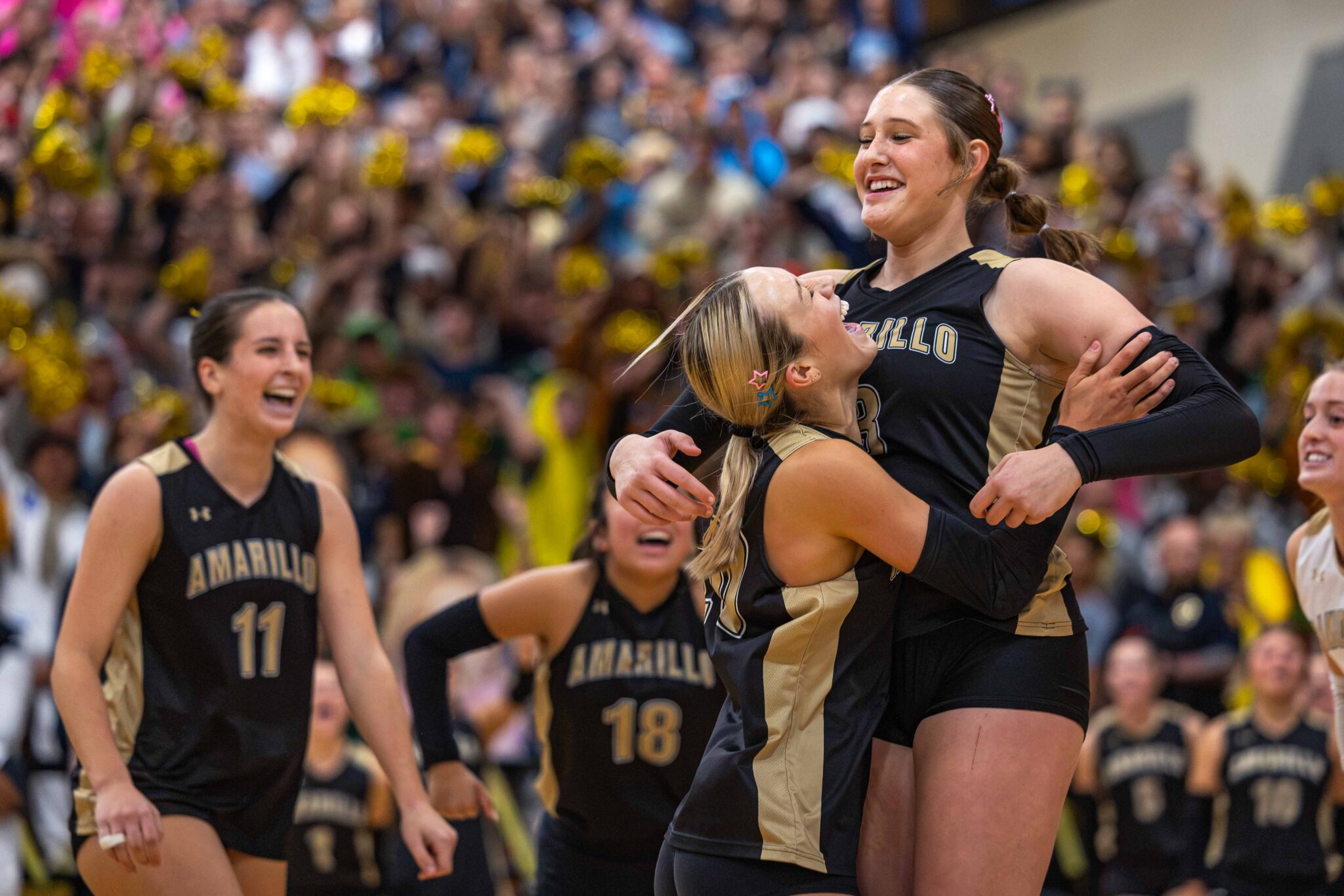 THS-AHS-VB-13 | Press Pass Sports Volleyball teammates in black and gold celebrate on court as a teammate is lifted up, crowd cheering behind them.