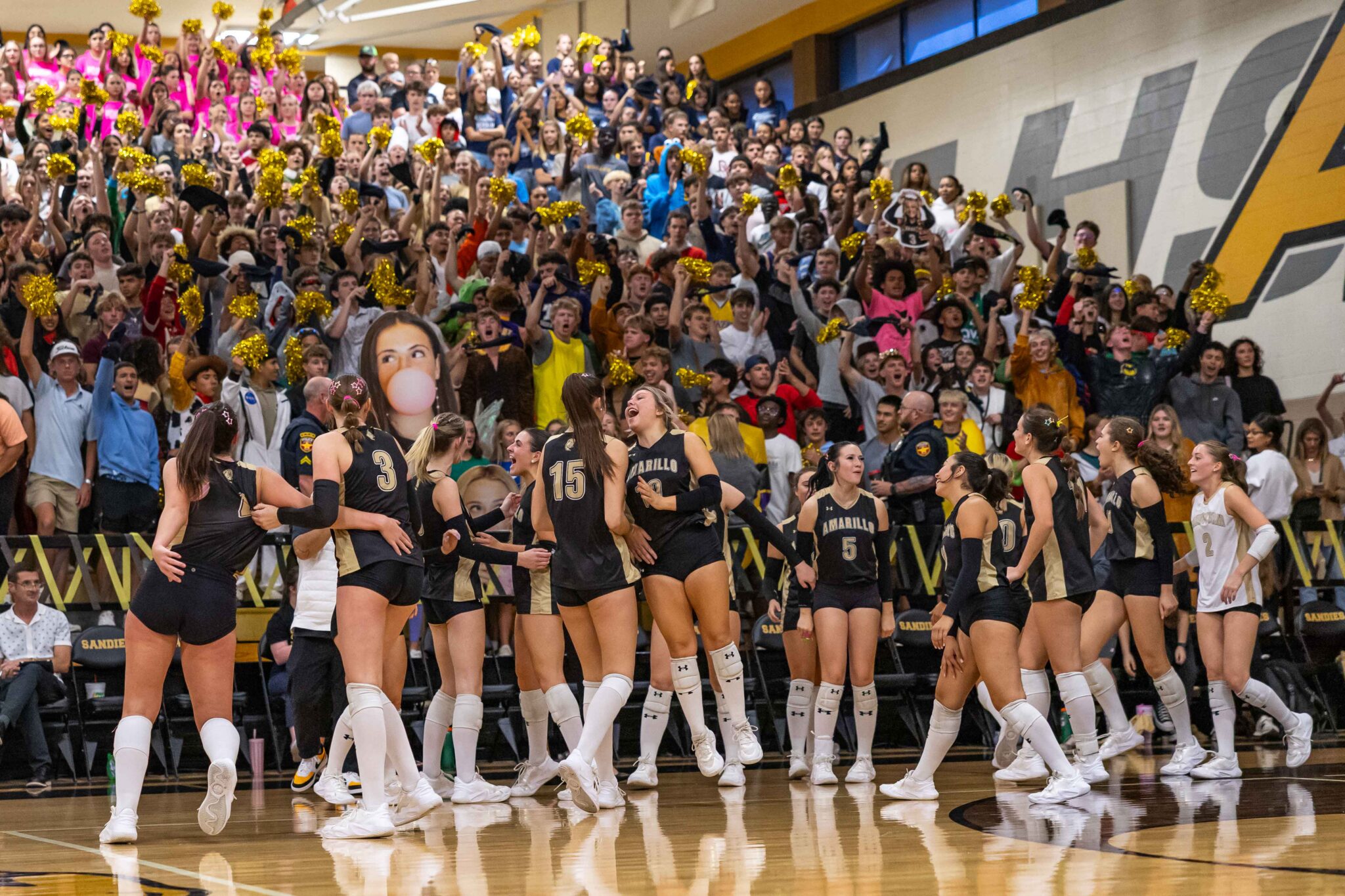 THS-AHS-VB-14 | Press Pass Sports Crowd of excited students in a gym cheering with gold pom-poms while basketball players in black uniforms stand in front on the court.