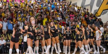 Crowd of excited students in a gym cheering with gold pom-poms while basketball players in black uniforms stand in front on the court.