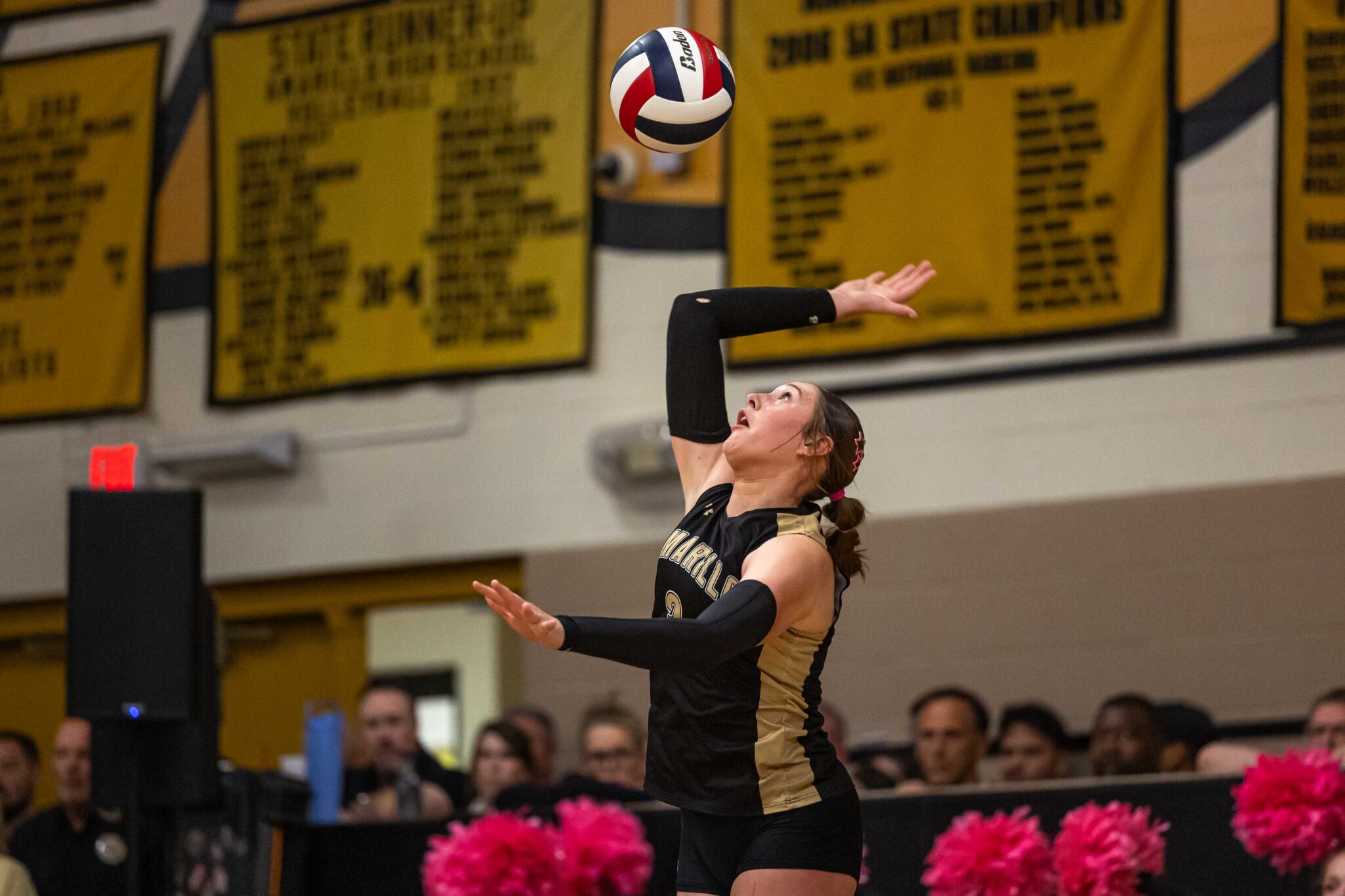 THS-AHS-VB-17 | Press Pass Sports Female volleyball player in black and gold uniform reaches up to set a ball during a match, gym banners in the background.