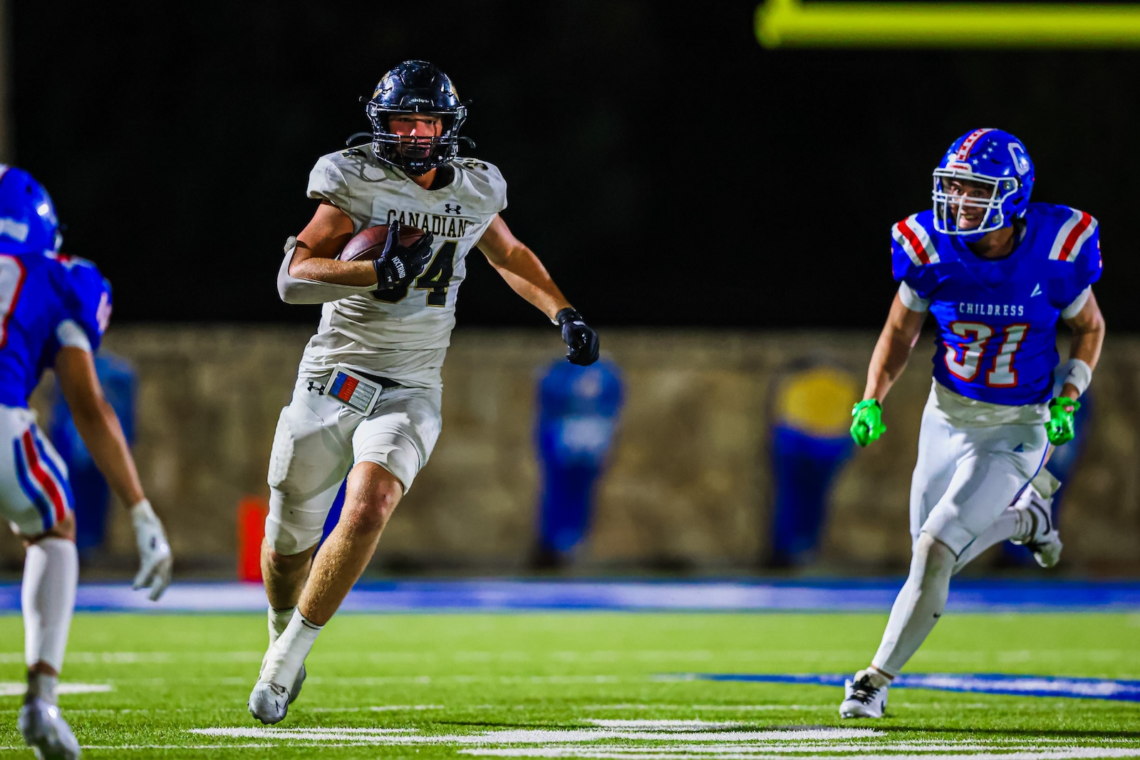 CT9A4515 | Press Pass Sports Canadian football player in white jersey carrying the ball, sprinting down the field as blue-uniform defenders chase him at night on a lit stadium turf