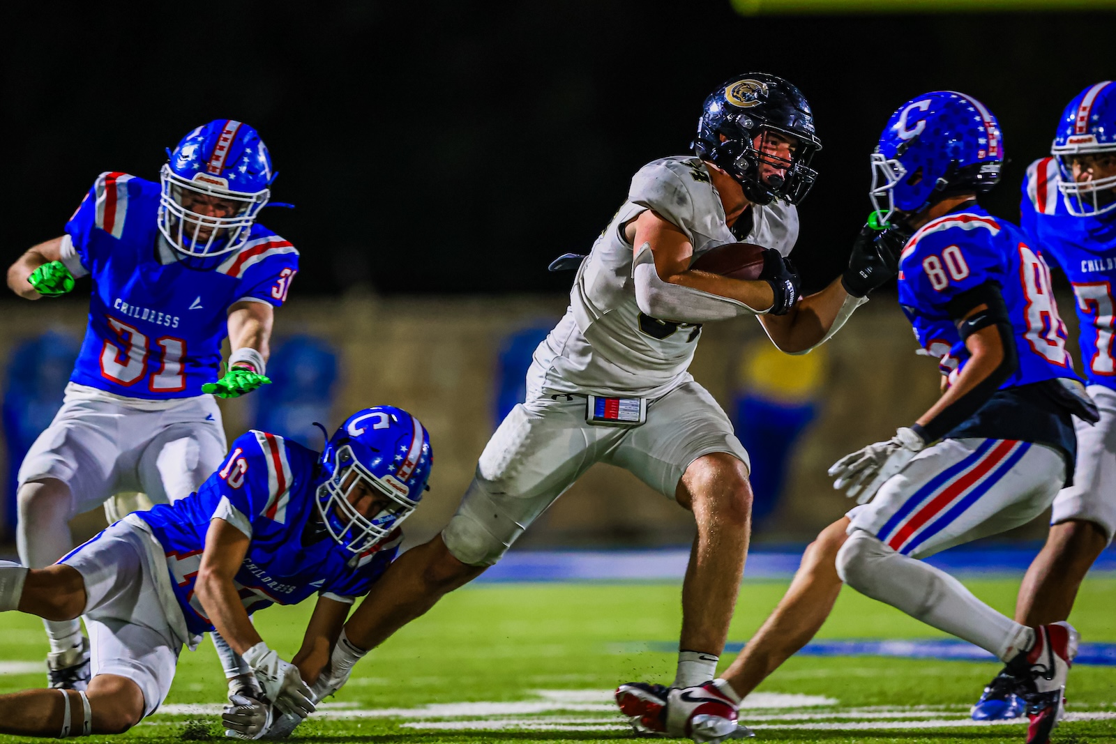 CT9A4552 | Press Pass Sports White-uniformed football player runs with the ball as blue-clad defenders close in during a night game on the field.