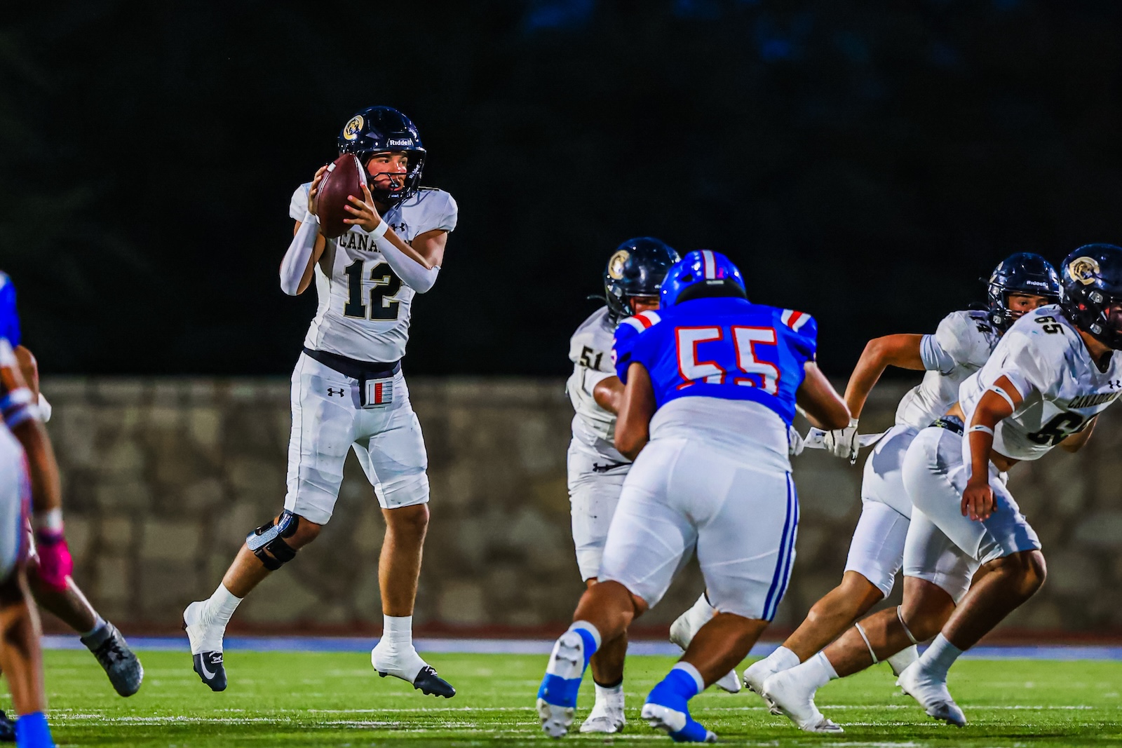 CT9A6526 | Press Pass Sports Quarterback in white #12 throws a pass during a nighttime football game as teammates and defenders move around him.