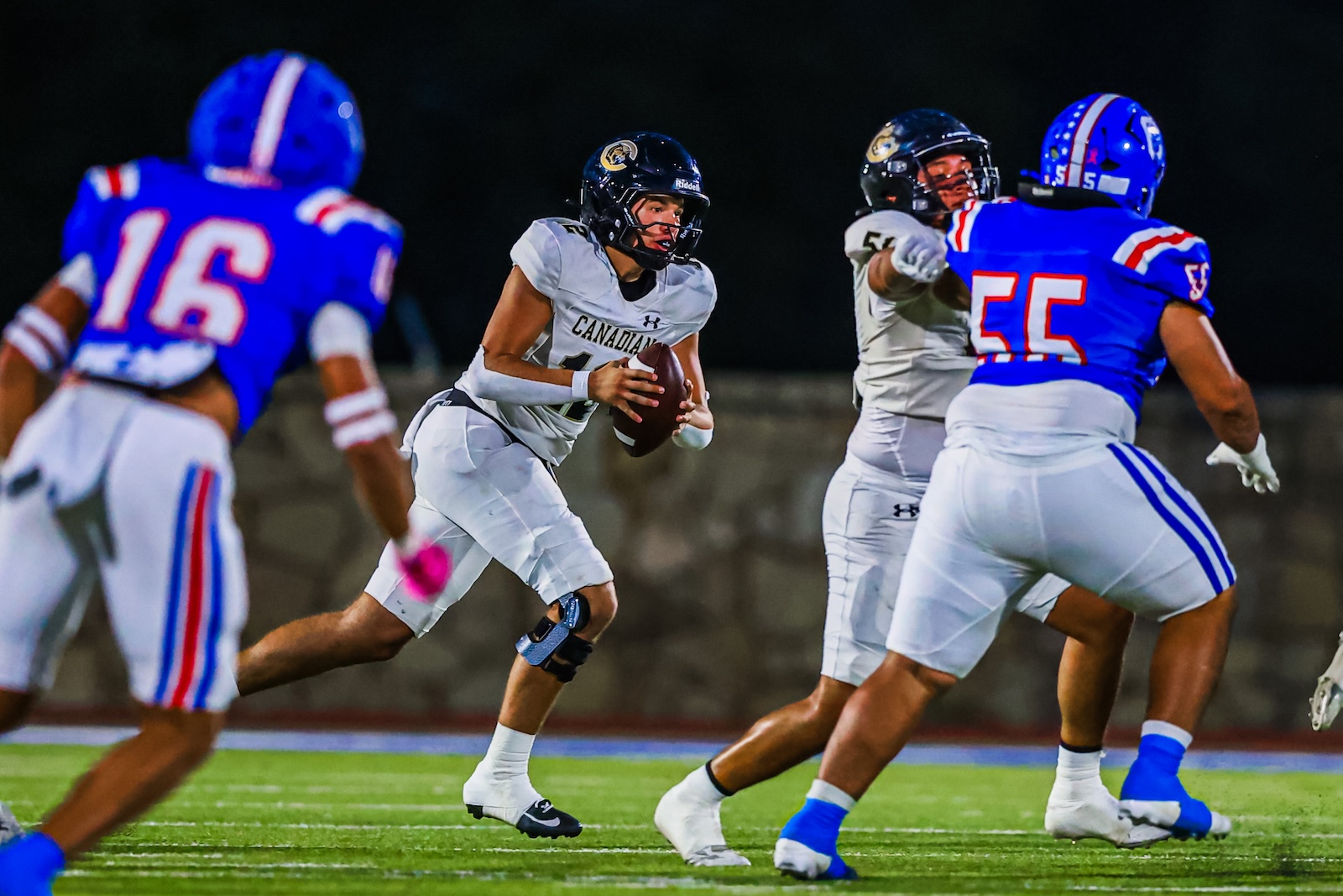 CT9A6552 | Press Pass Sports Quarterback in white Canadian uniform runs with the ball while blue-clad defenders close in during a night game.