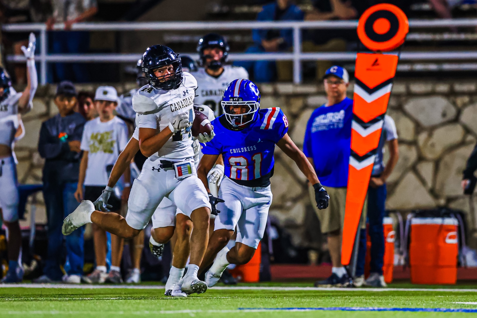CT9A6822 | Press Pass Sports White-uniform ball carrier runs with the ball as a blue-uniform defender pursues on a football field near the sideline pylon.