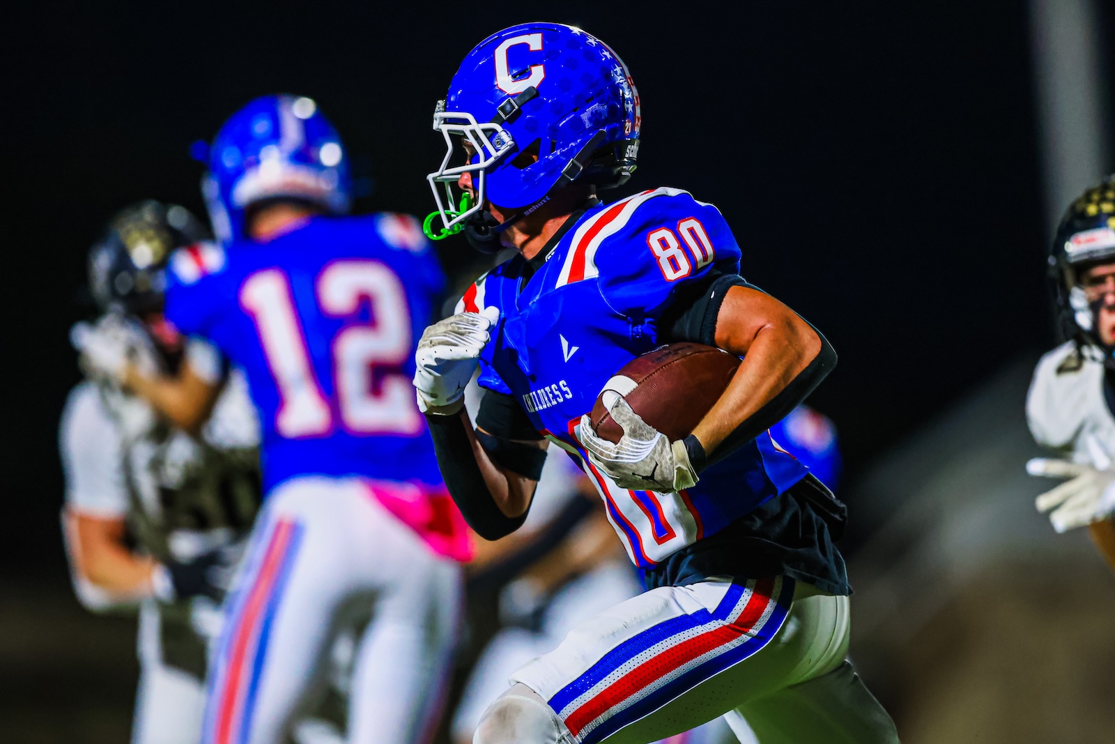 CT9A7731 | Press Pass Sports Football player in a blue uniform clutching the ball and running forward during a nighttime game.